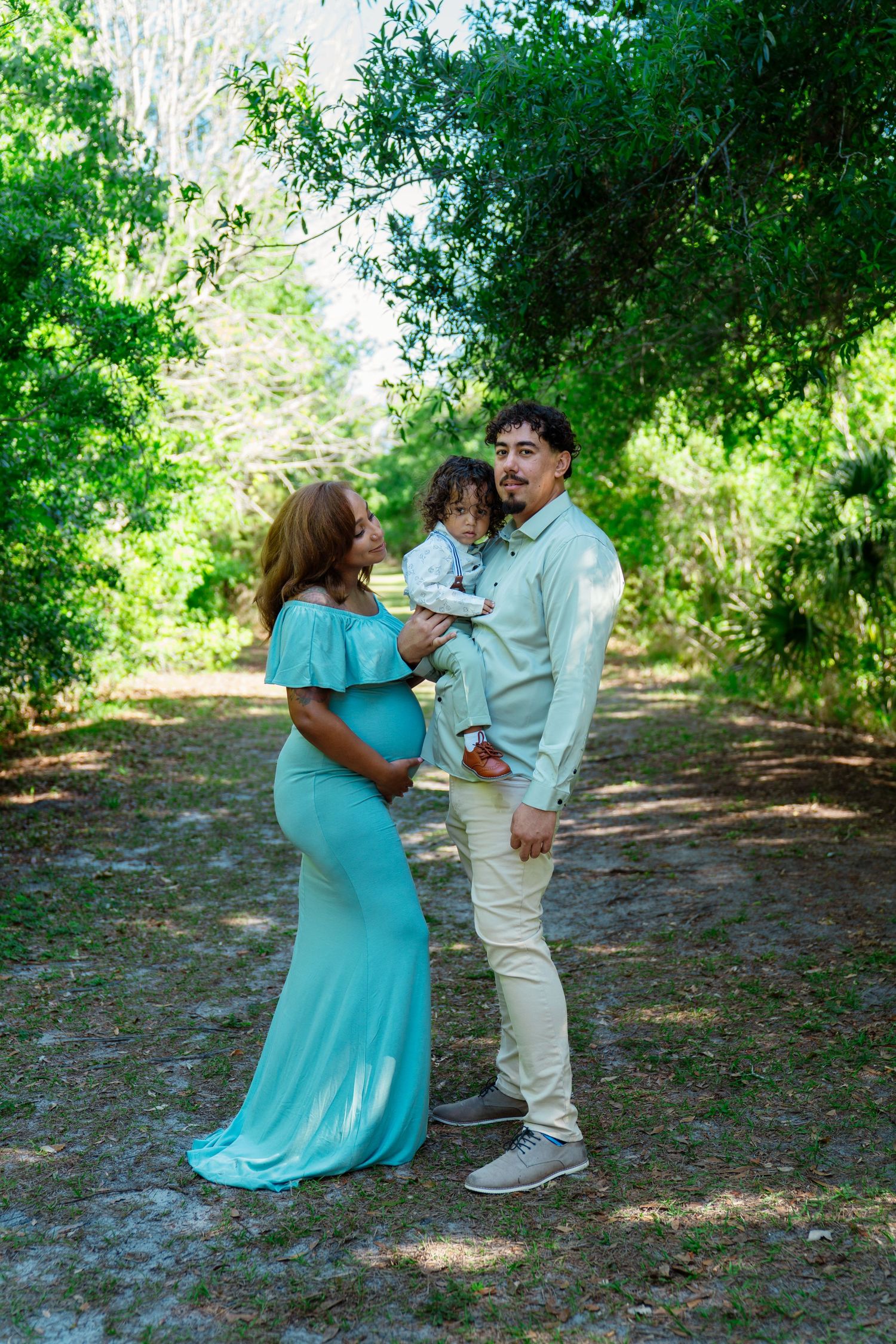 Pregnant woman in teal gown with husband holding toddler during outdoor maternity photo session.