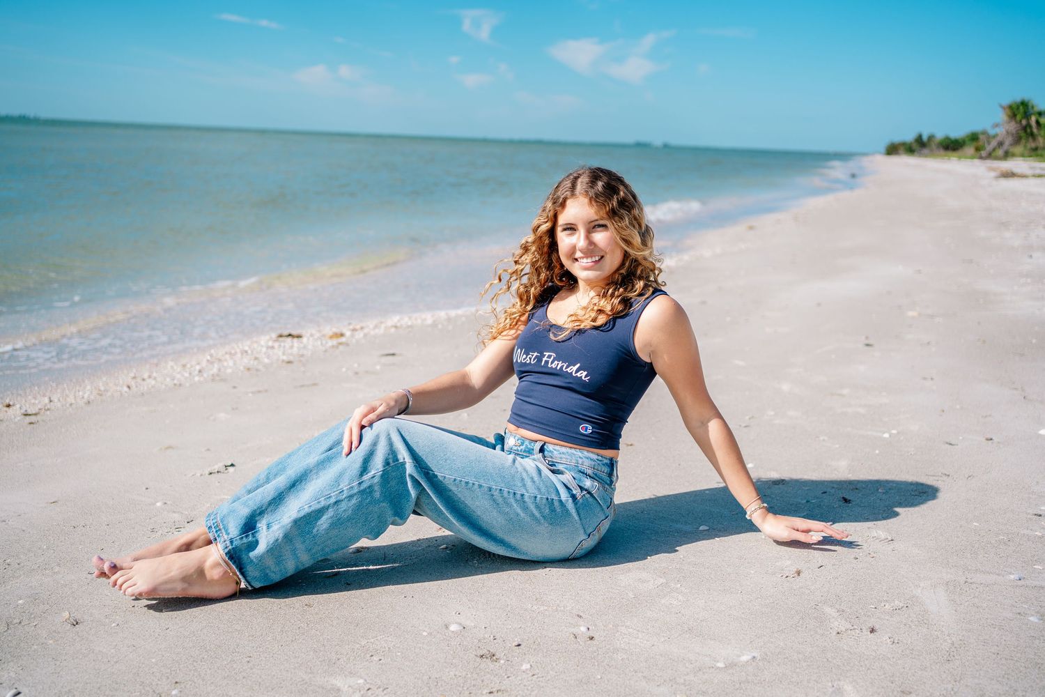 Young woman with curly hair sitting on a sunny white sand beach wearing jeans and a navy tank top.