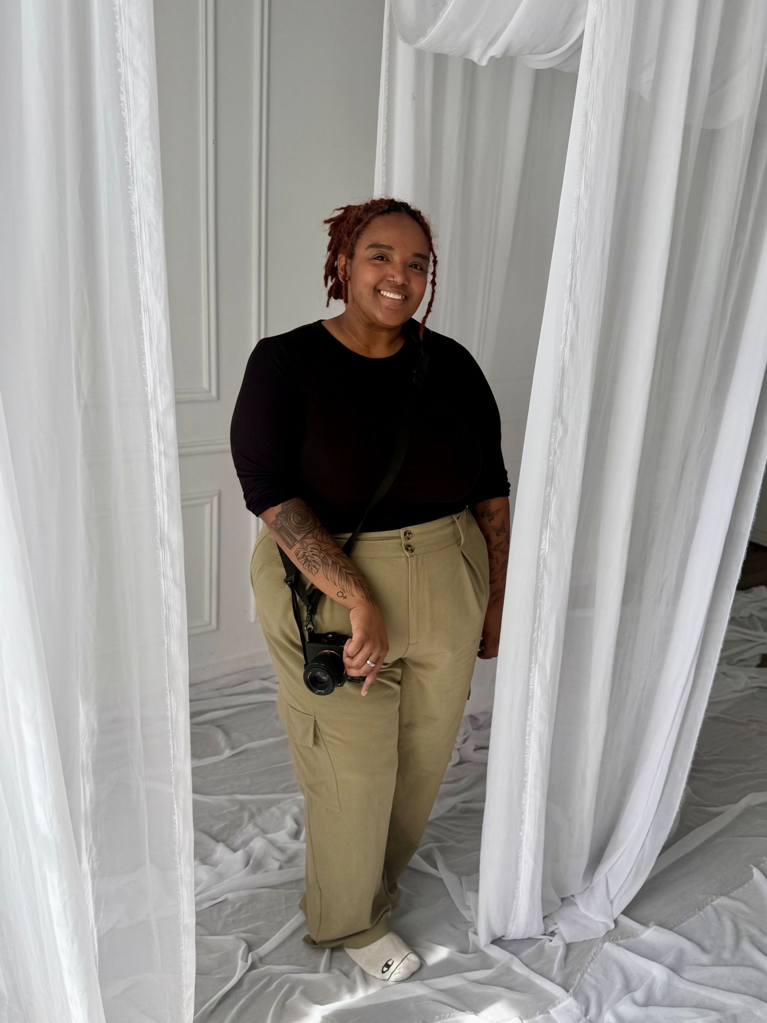 Smiling person in black top and khaki cargo pants standing between white curtains holding a camera.