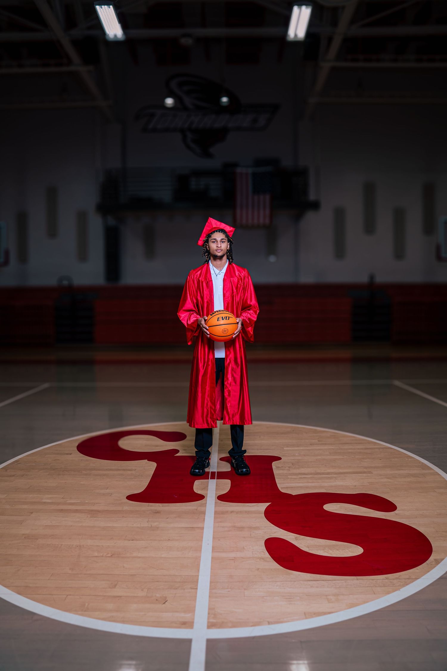 Graduate in red cap and gown holds basketball on school gym court center logo.