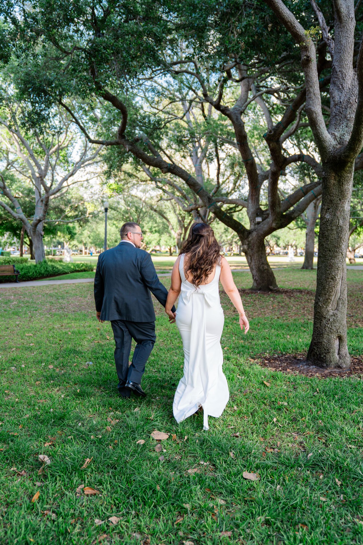 Bride and groom walk hand in hand through a lush green park, bride in white gown, groom in dark suit.