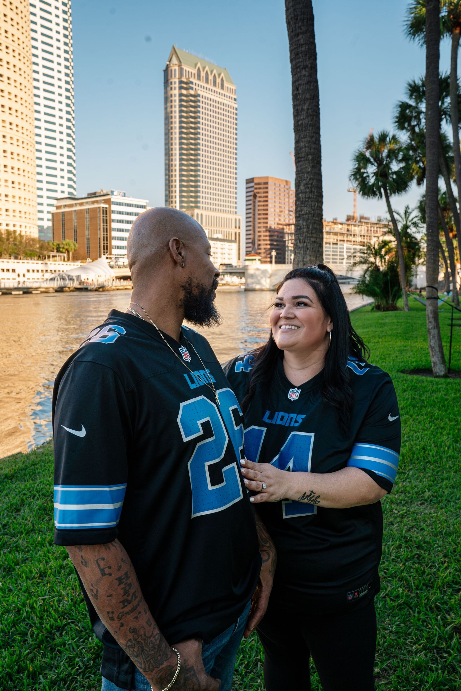 Couple wearing matching Detroit Lions NFL jerseys smiling at each other by Tampa Bay waterfront skyline.