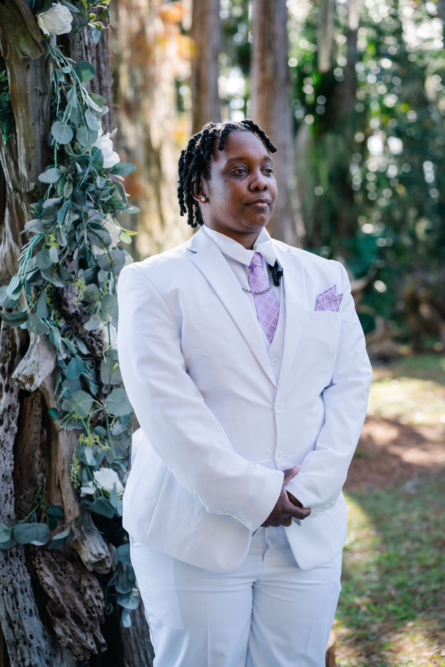 Person in white suit with lavender tie and pocket square stands outdoors near floral-decorated tree at wedding ceremony.