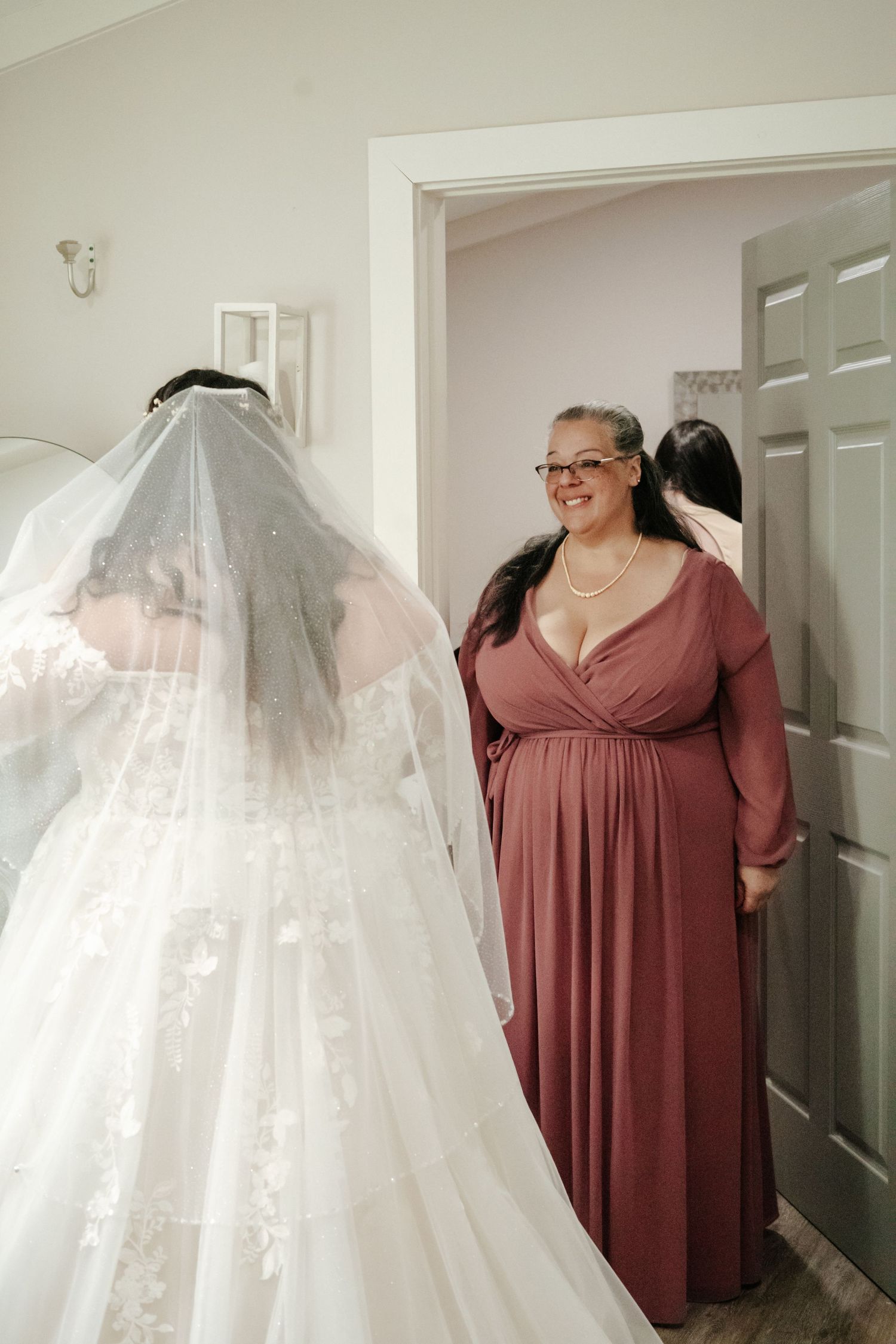 White wedding dress hanging beside a person wearing a mauve dress in a bright room.
