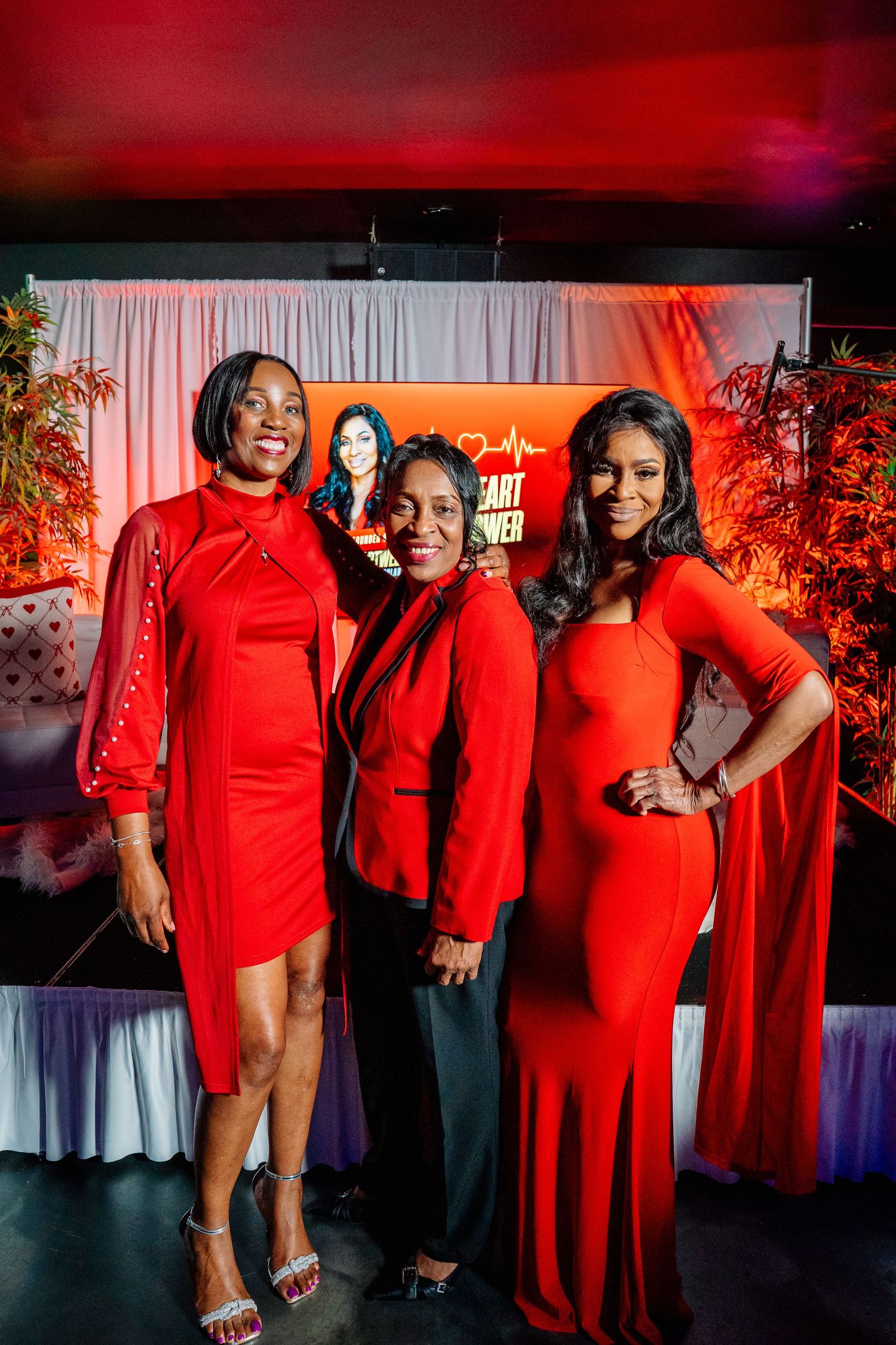Three women in red outfits pose together at a Heart Power event with decorative backdrop.