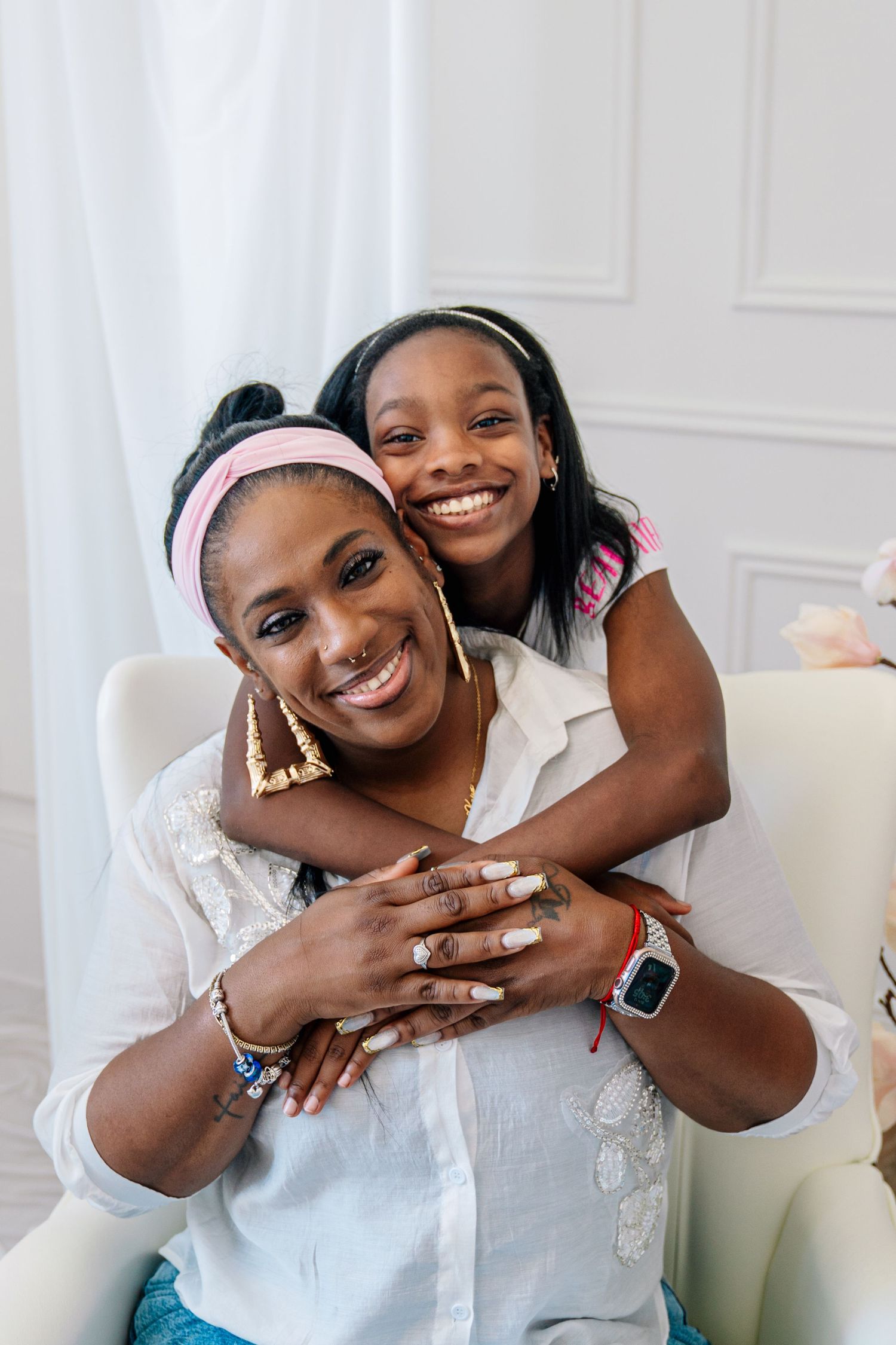 A young girl hugs a smiling woman from behind as they pose together in a bright white room.