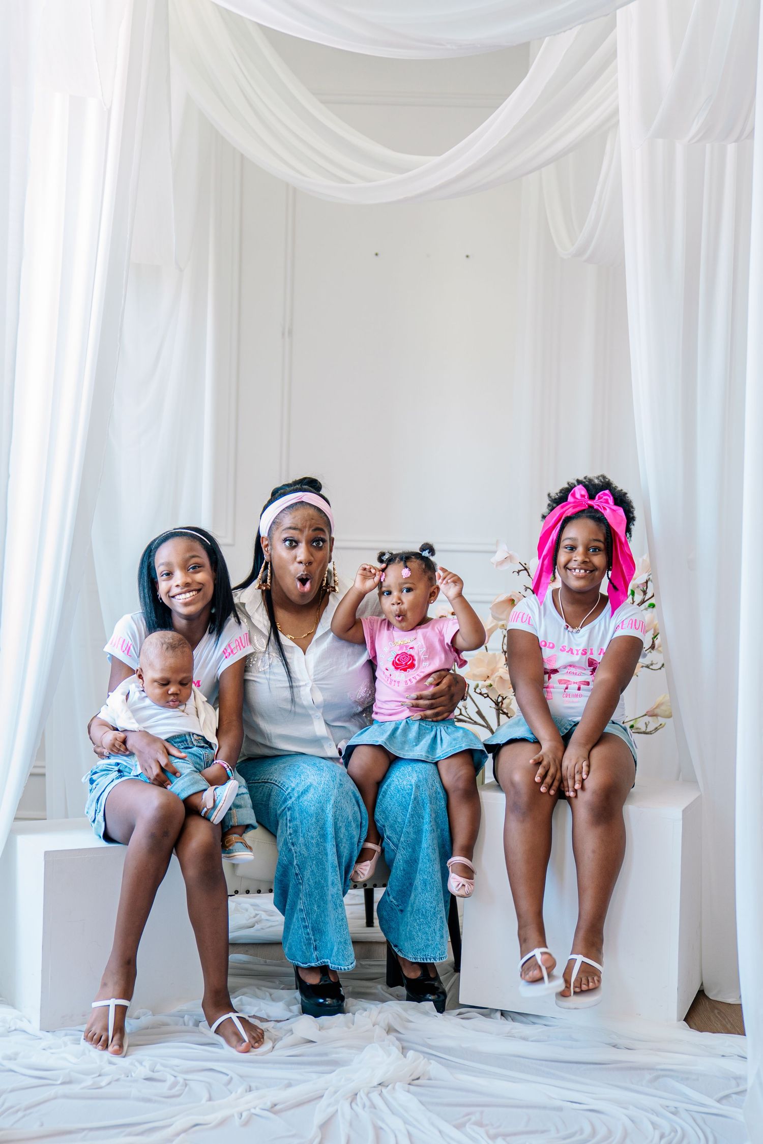 A woman and three girls pose together on a white bench, dressed in matching pink and white outfits, with a baby.