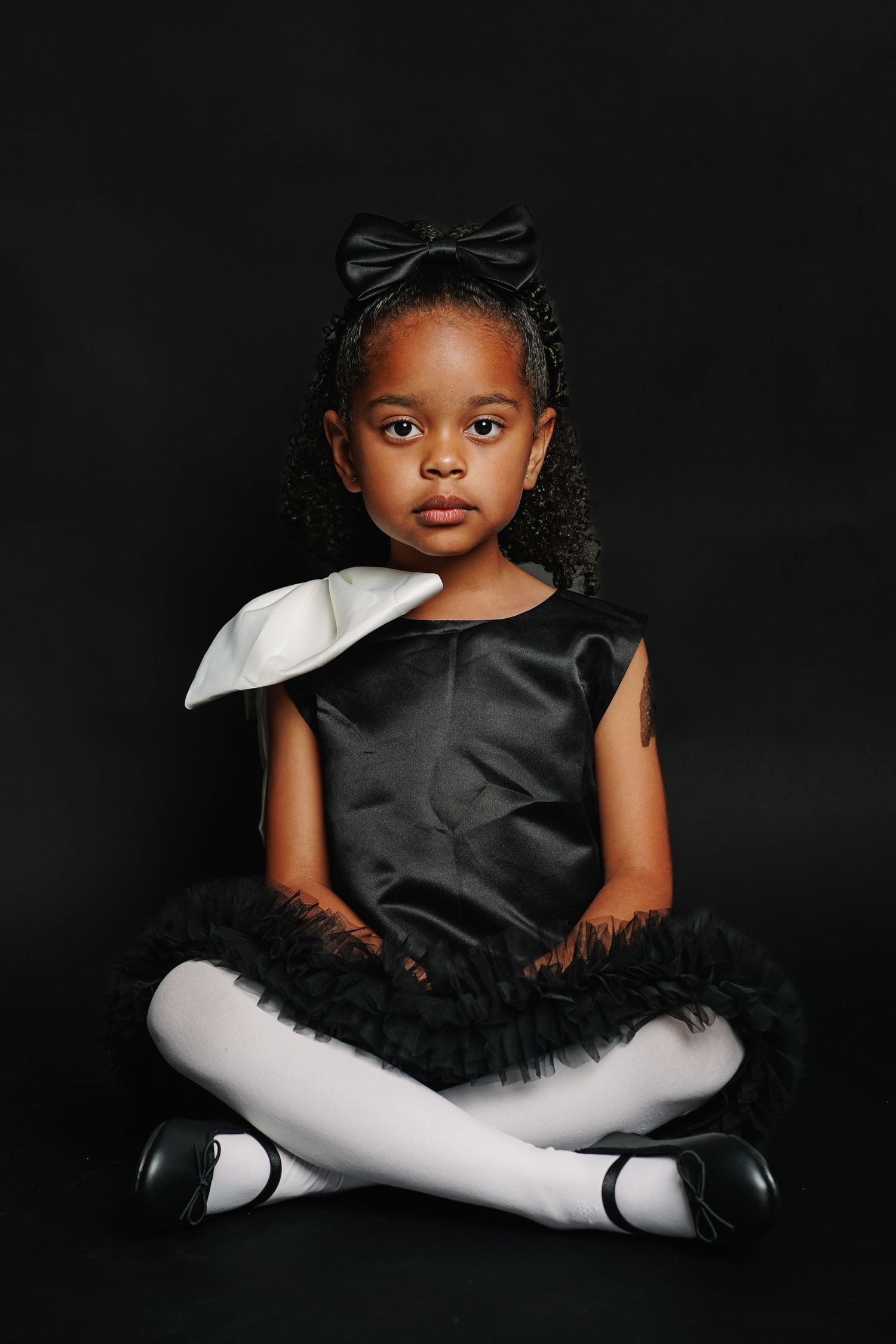 Young Black girl in black satin dress with white bow and tutu skirt, white tights, sitting cross-legged on dark background.