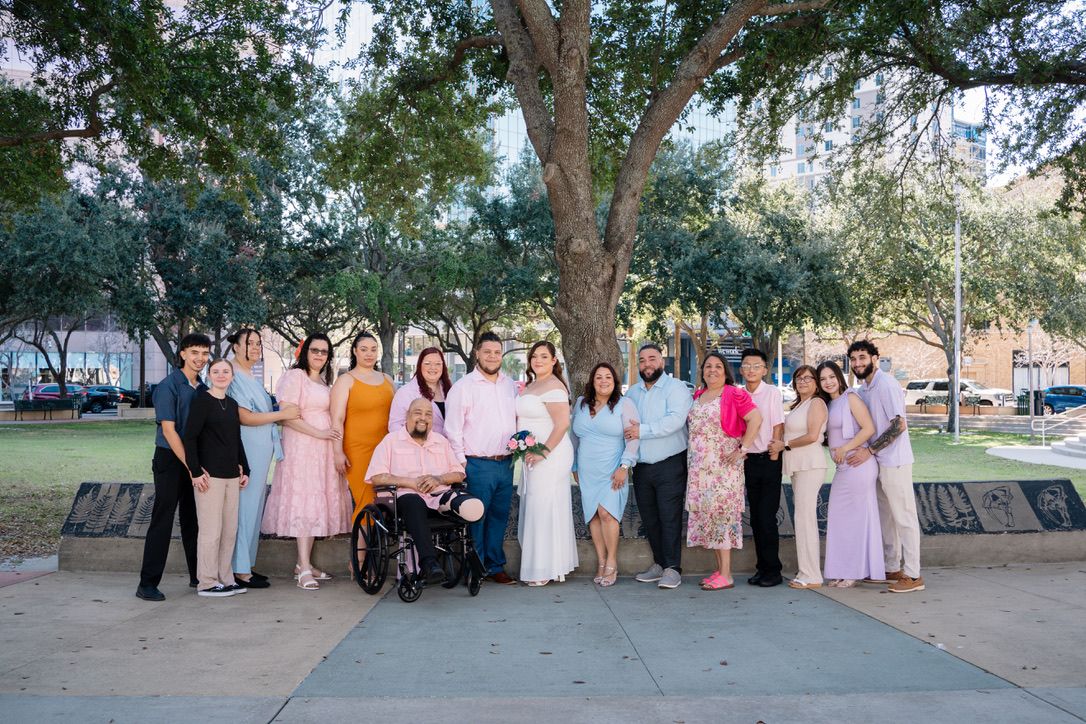 A group in formal attire gathers under a large tree for a special occasion photo in a park setting.