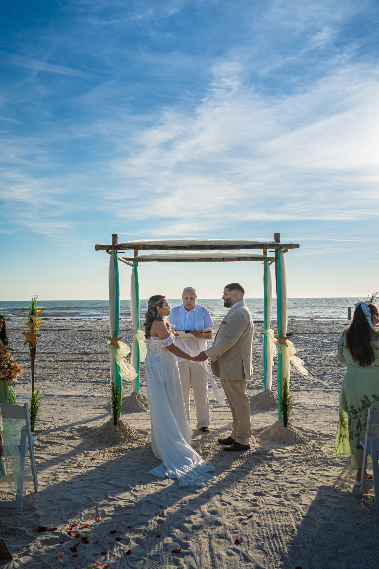 A beachside wedding ceremony takes place under a bamboo arch with the ocean and blue sky as a backdrop.