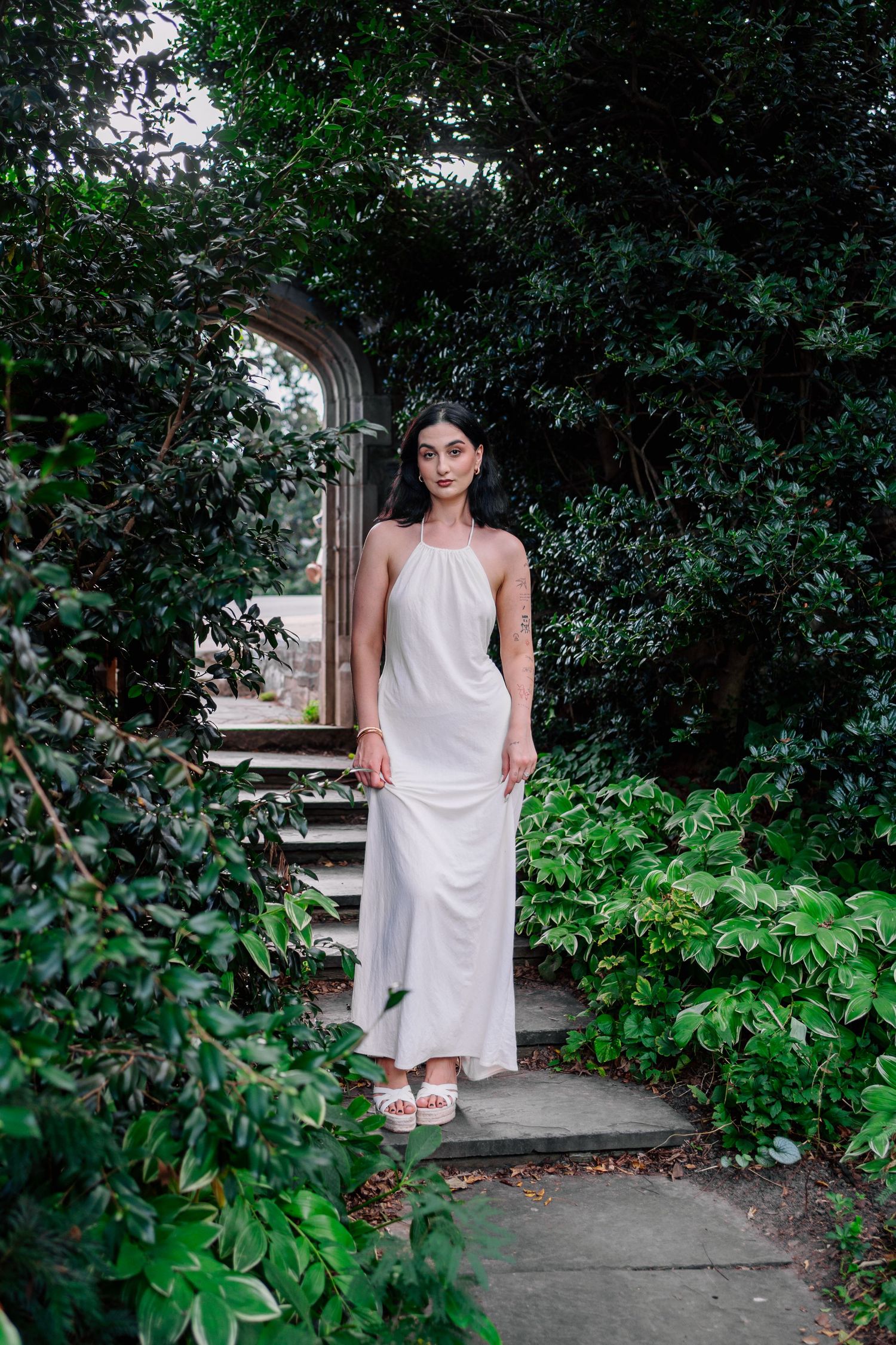Woman in white halter maxi dress standing on stone path surrounded by lush green garden archway.