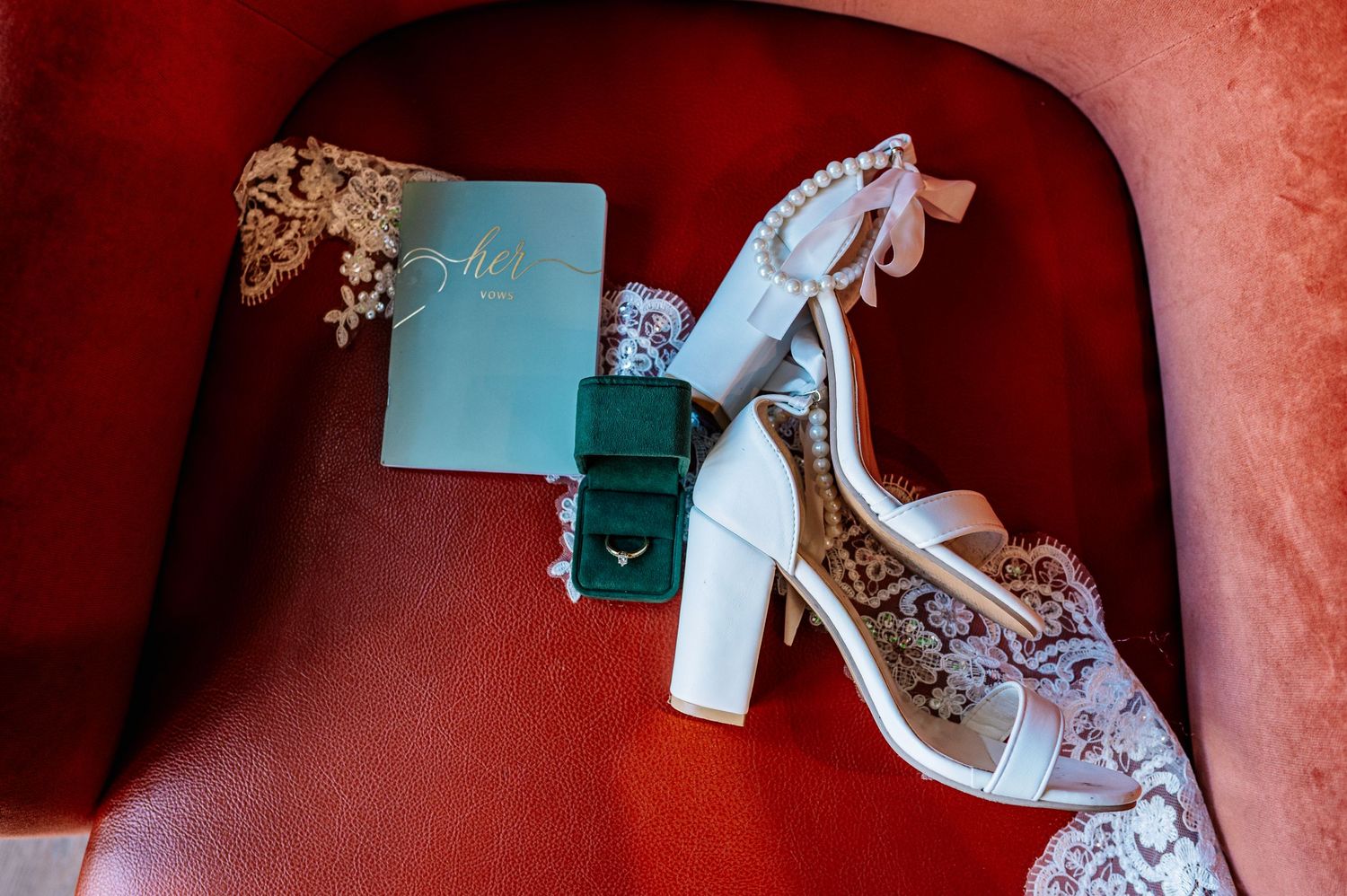 White bridal heels with pearl ankle strap beside a green ring box and lace veil on a red velvet chair.