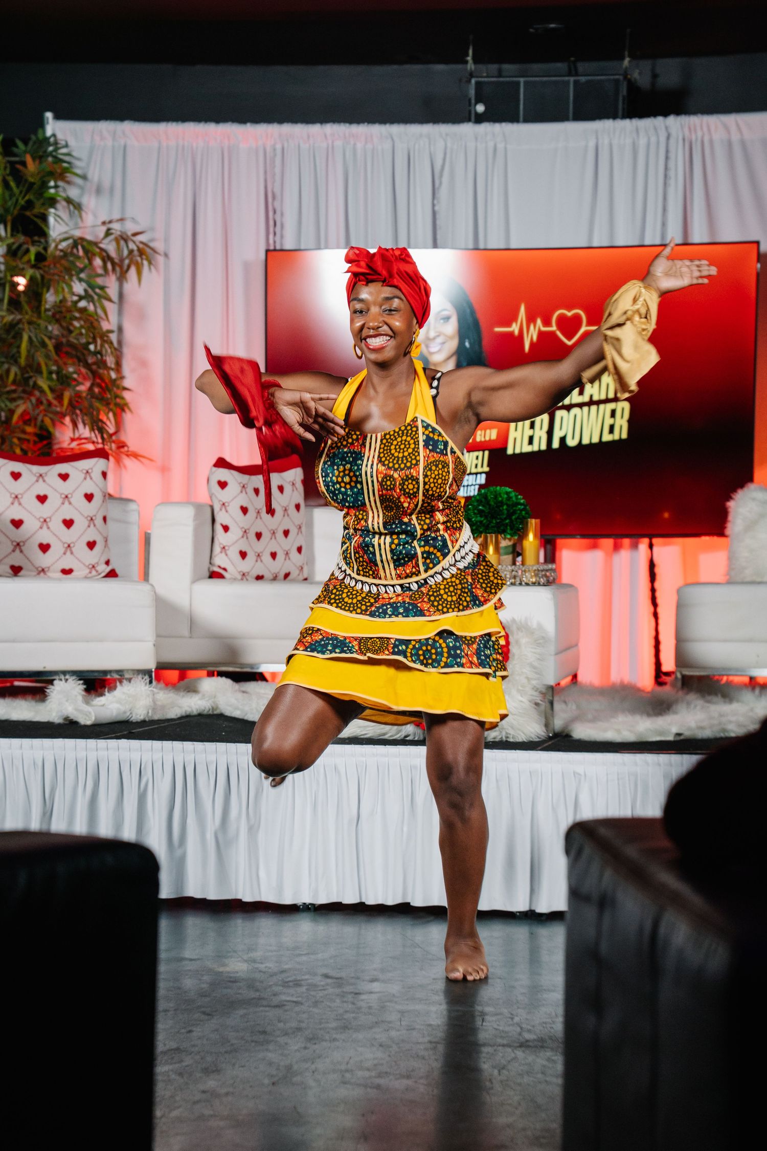 Woman in colorful African print dress and red headwrap dancing energetically on stage at a cultural event.