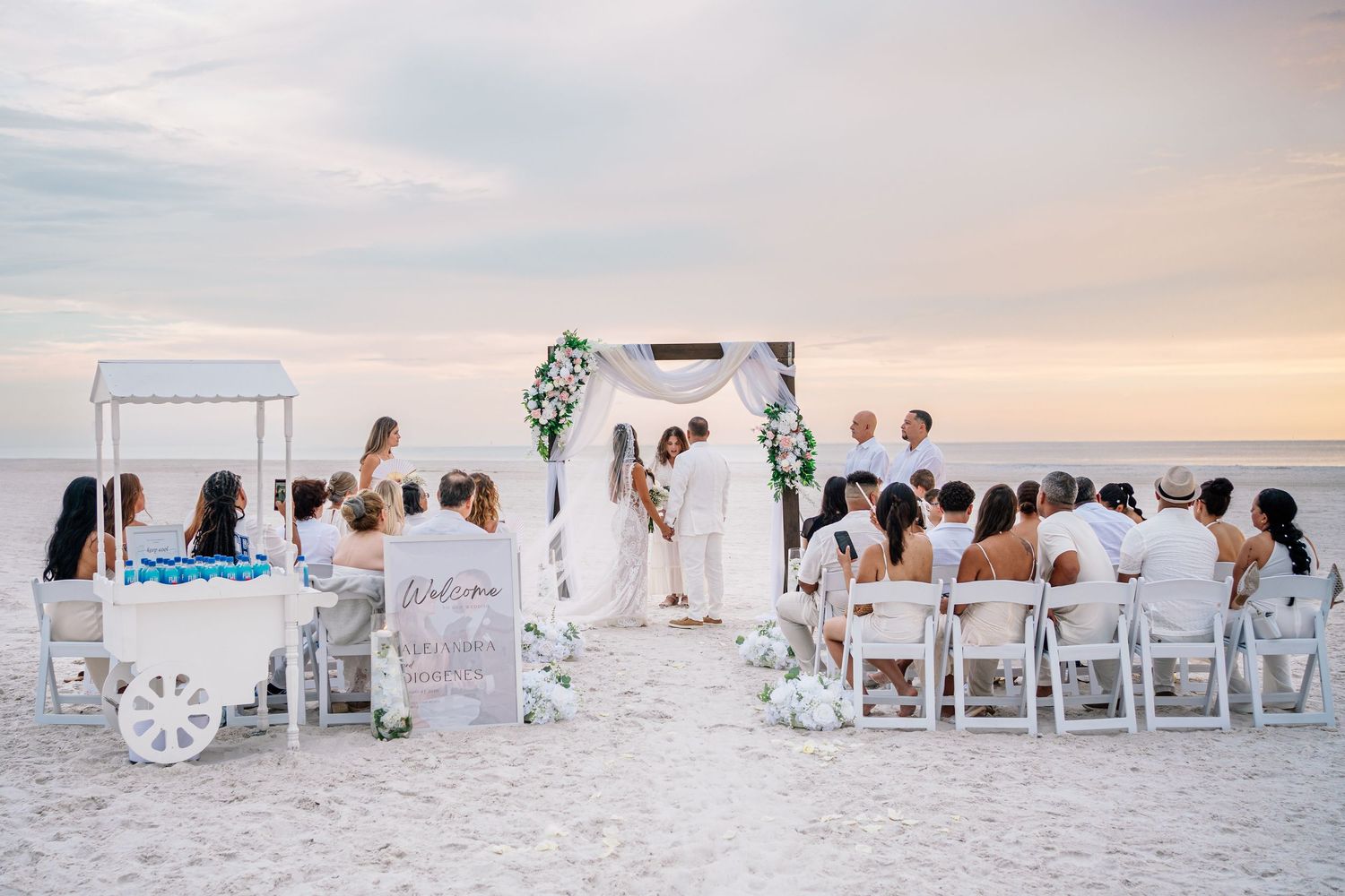 Romantic beach wedding ceremony at sunset with guests seated on white chairs facing couple at floral arch.