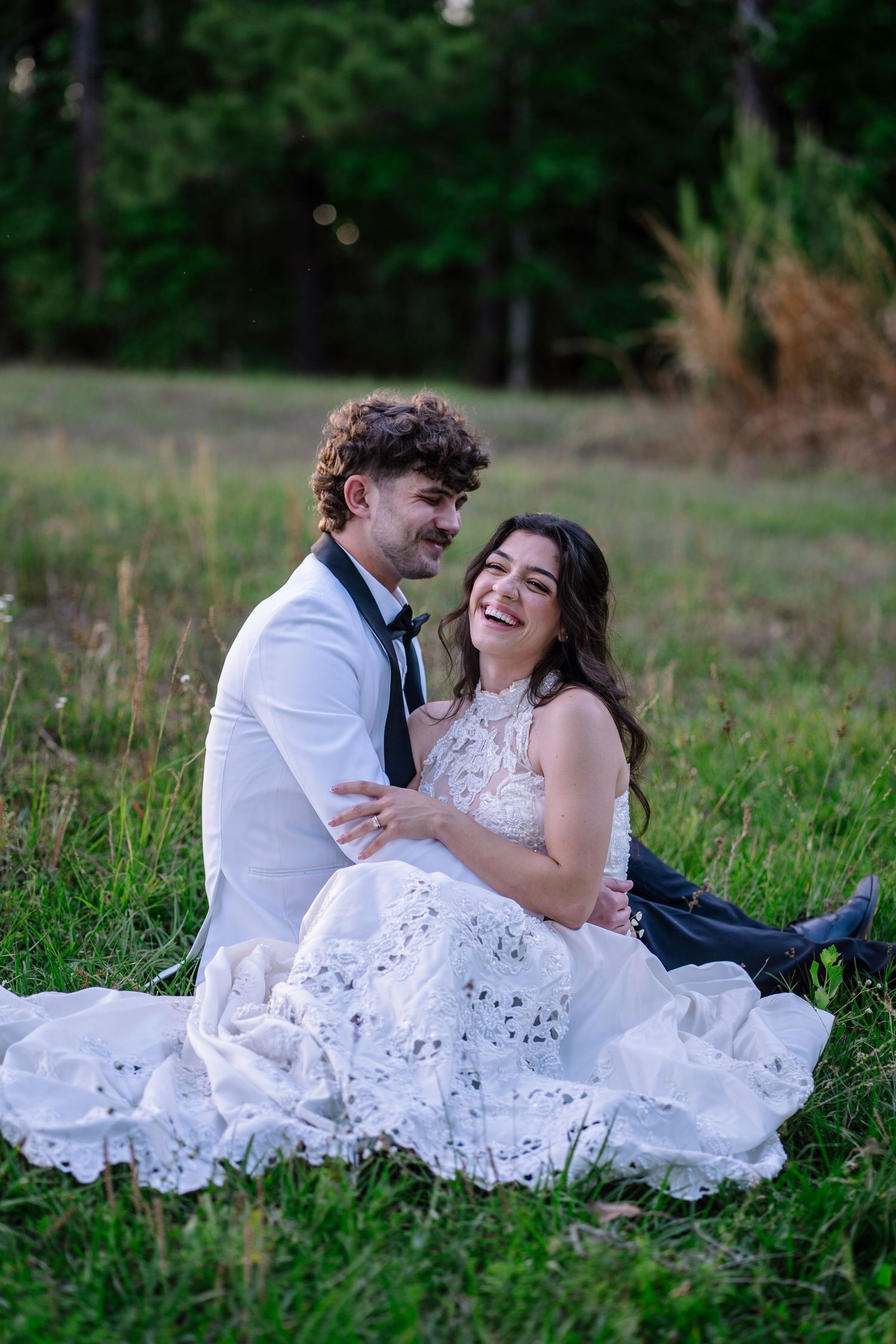A joyful bride and groom sit together on green grass, laughing and embracing in an outdoor wedding photo.