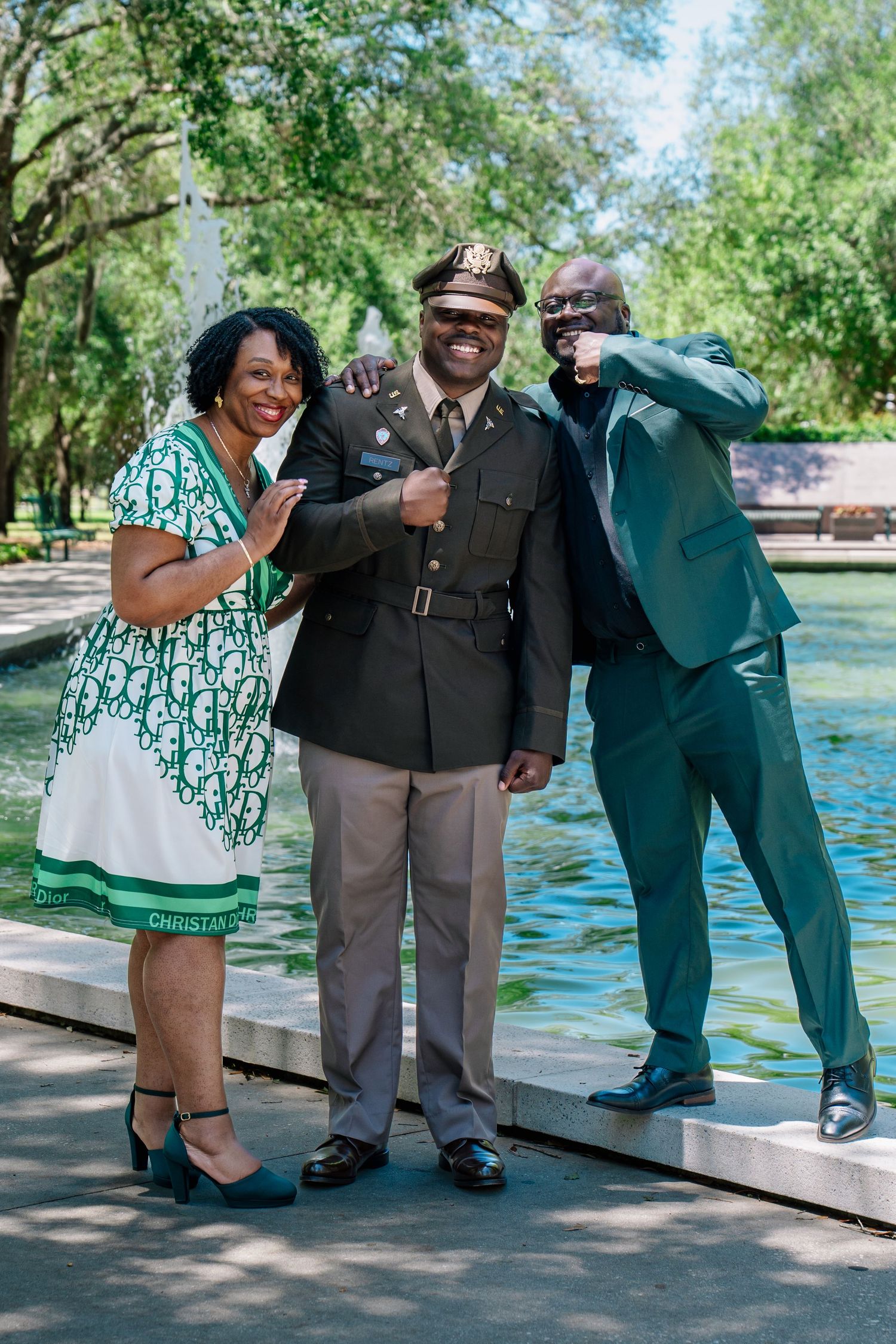 Military officer in uniform celebrates with two proud family members outdoors near a fountain on a sunny day.