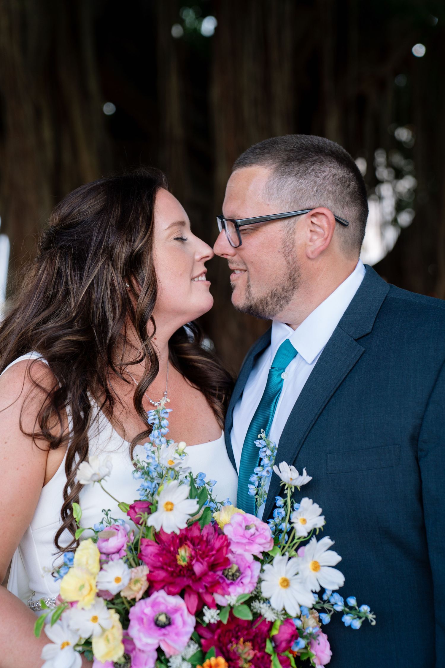 A bride and groom share a romantic nose-to-nose moment, with a vibrant colorful bouquet in the foreground.