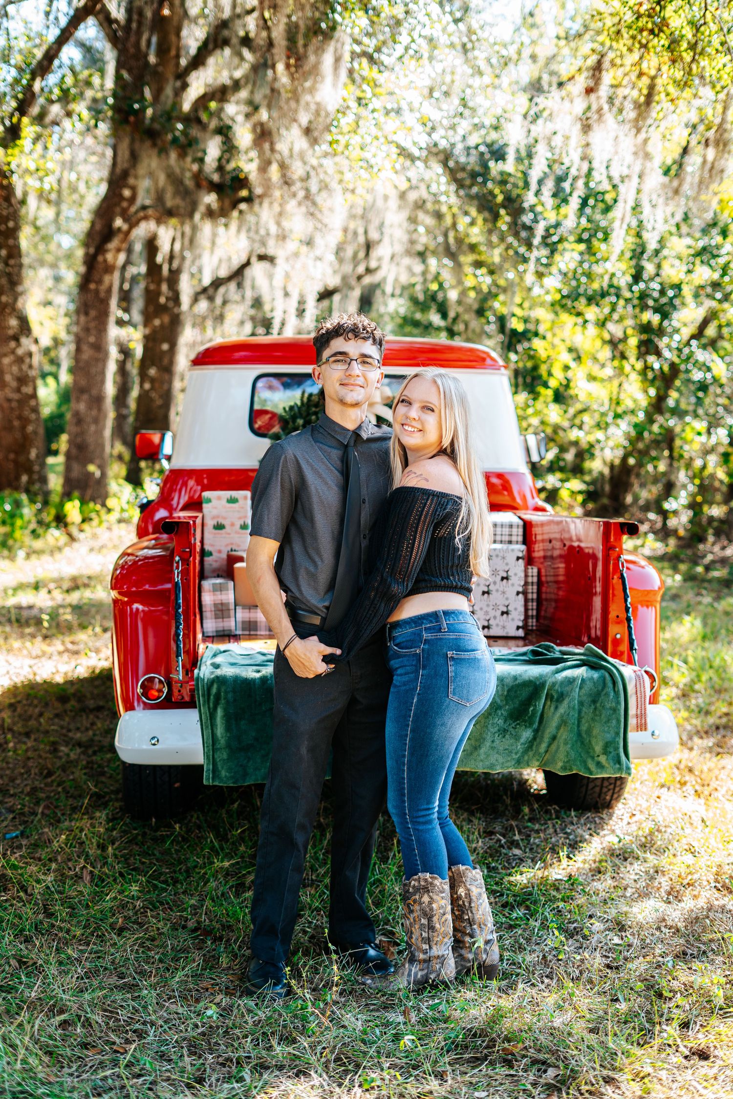 A couple shares romantic moments by a vintage red pickup truck in a sunlit wooded setting.