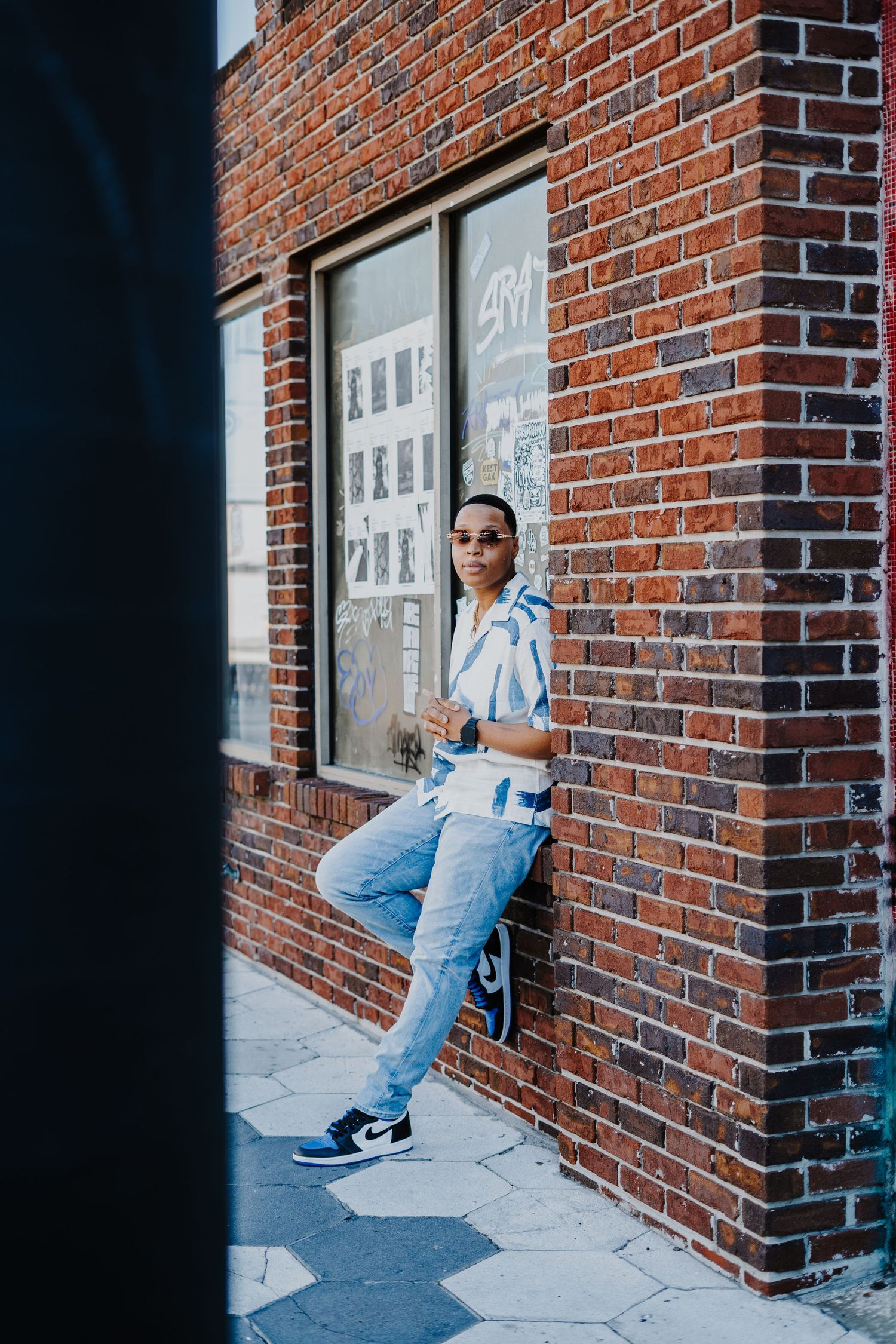 Person in denim overalls and sneakers sits against red brick wall in urban setting.