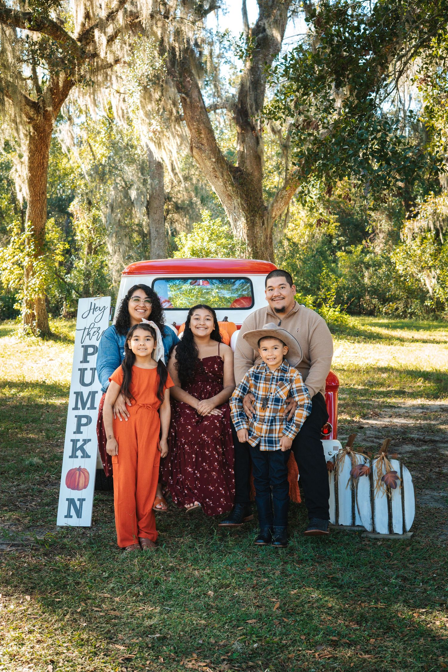 Family poses for Christmas photos with vintage red truck and holiday decorations in wooded setting.
