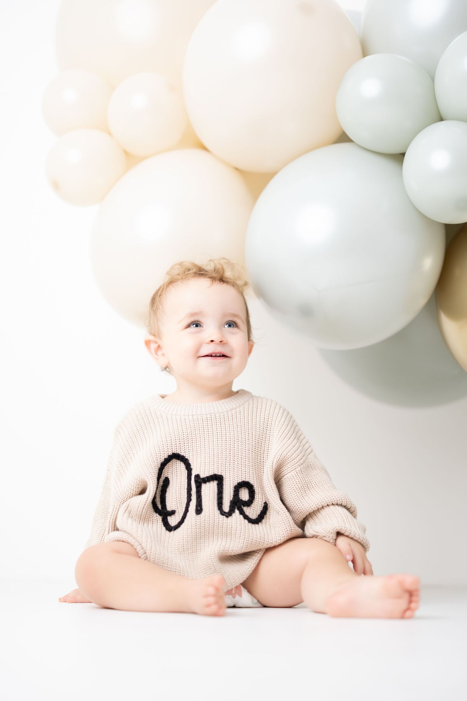 Smiling baby wearing a beige 'One' sweater sits barefoot in front of white and sage green balloons for first birthday.