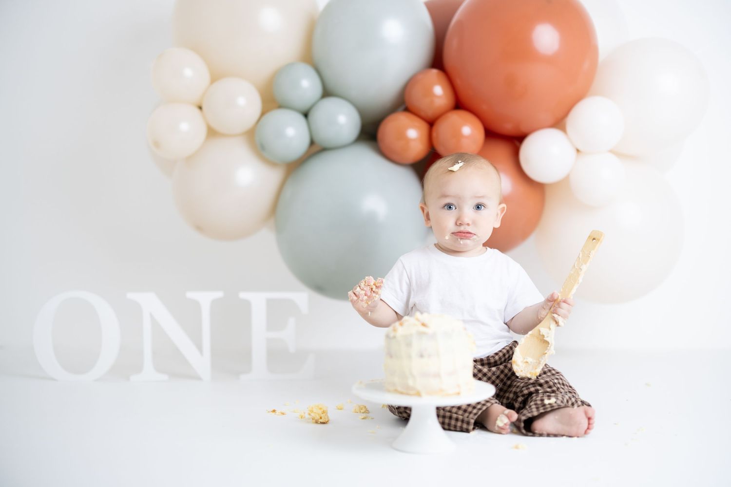 Baby sitting on white backdrop with pastel pink, gray and white balloon arch decorations for first birthday photoshoot.