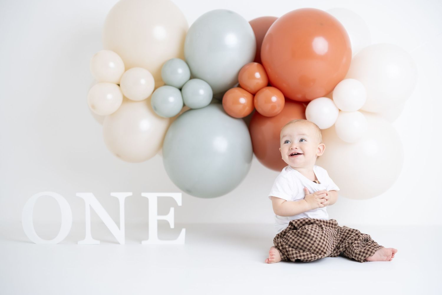Baby lies on floor surrounded by soft pastel blue, coral and white balloon garland for first birthday photoshoot.