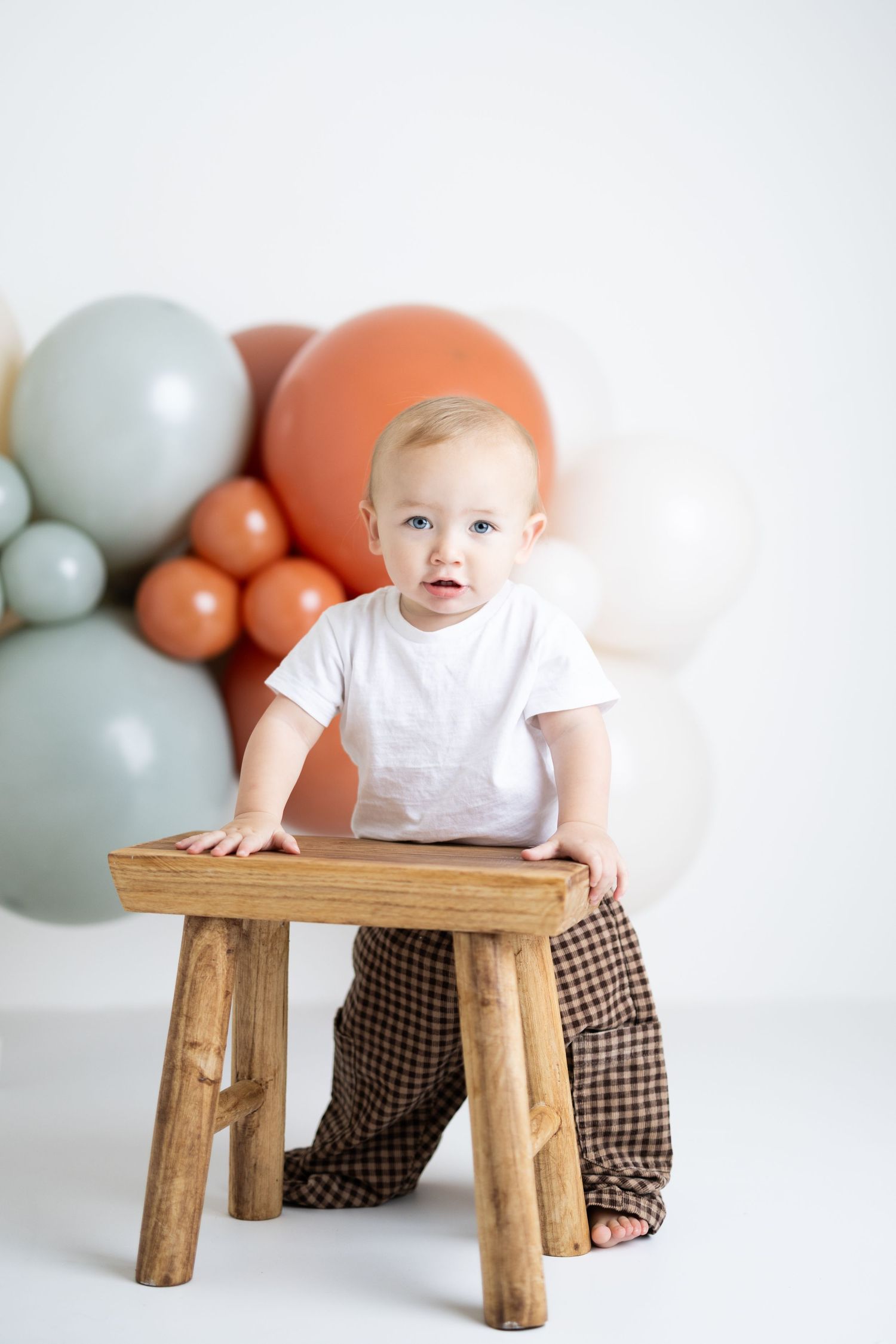 Baby in white shirt and plaid pants standing at wooden stool with colorful balloons in background.