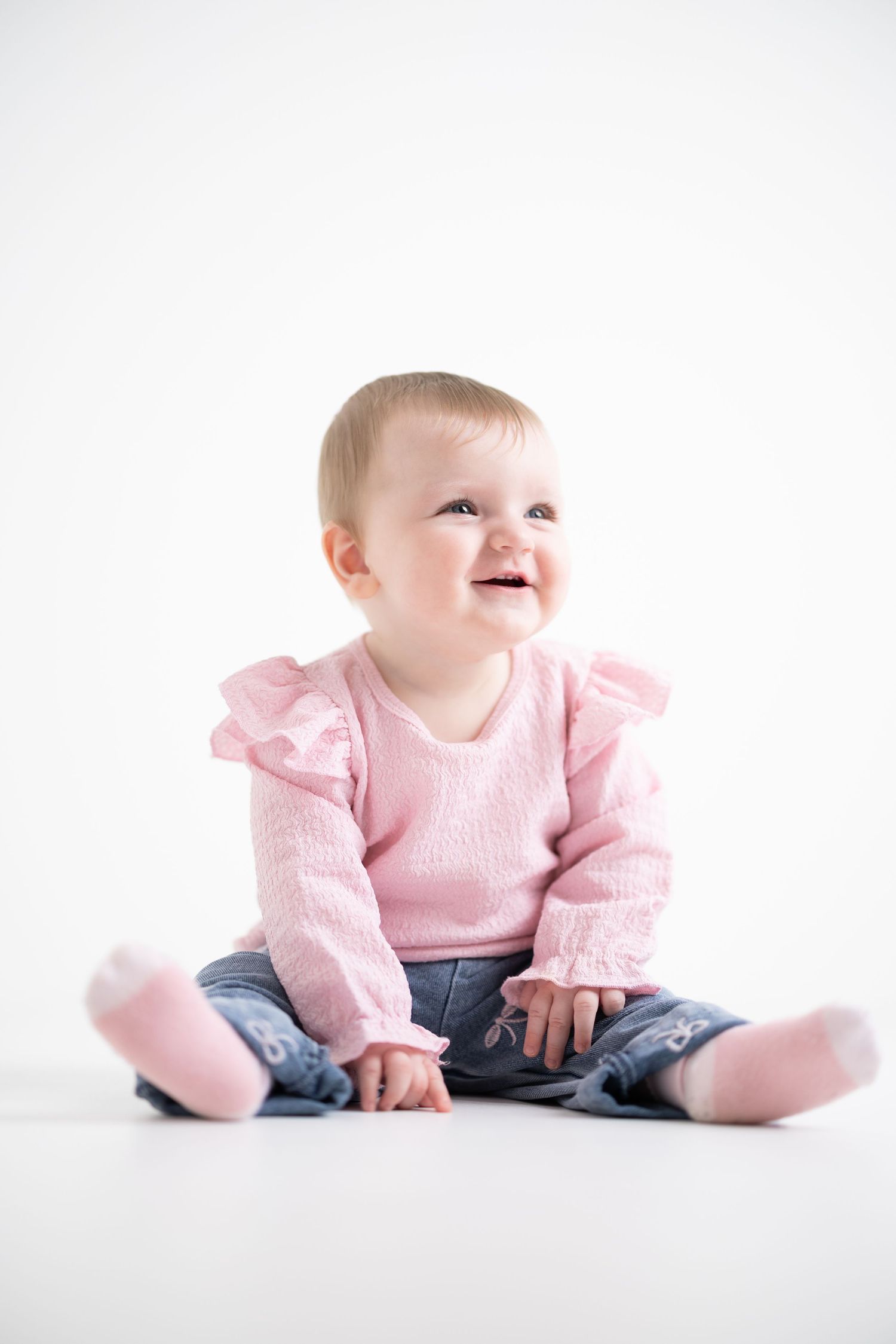 Baby wearing pink sweater and jeans sits on white background while smiling and looking up.
