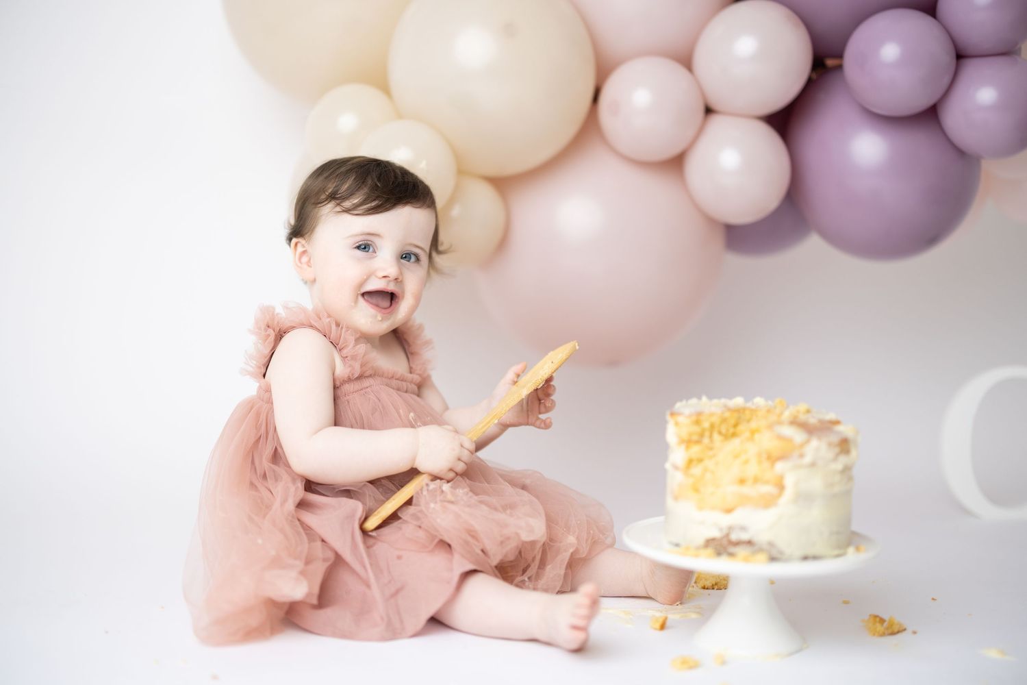 Baby in pink dress sits with birthday cake and balloons during cake smash photo session with purple and pink decor.