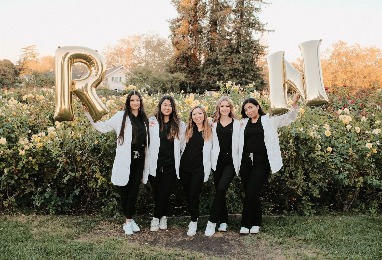 Group of graduates in white coats posing in front of large gold RN letters outdoors.