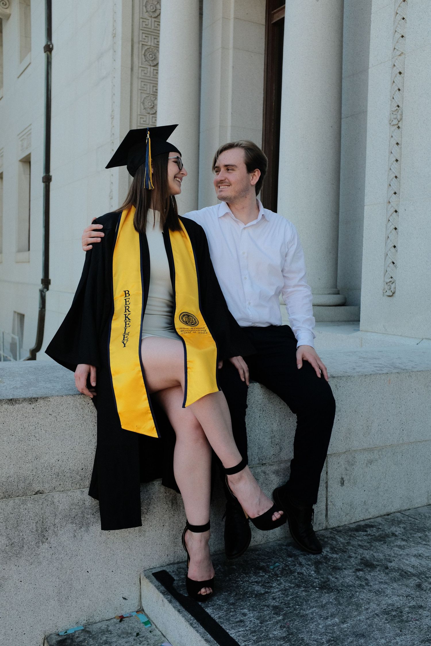 Graduate in cap and gown sits with companion on stone steps of columned building.
