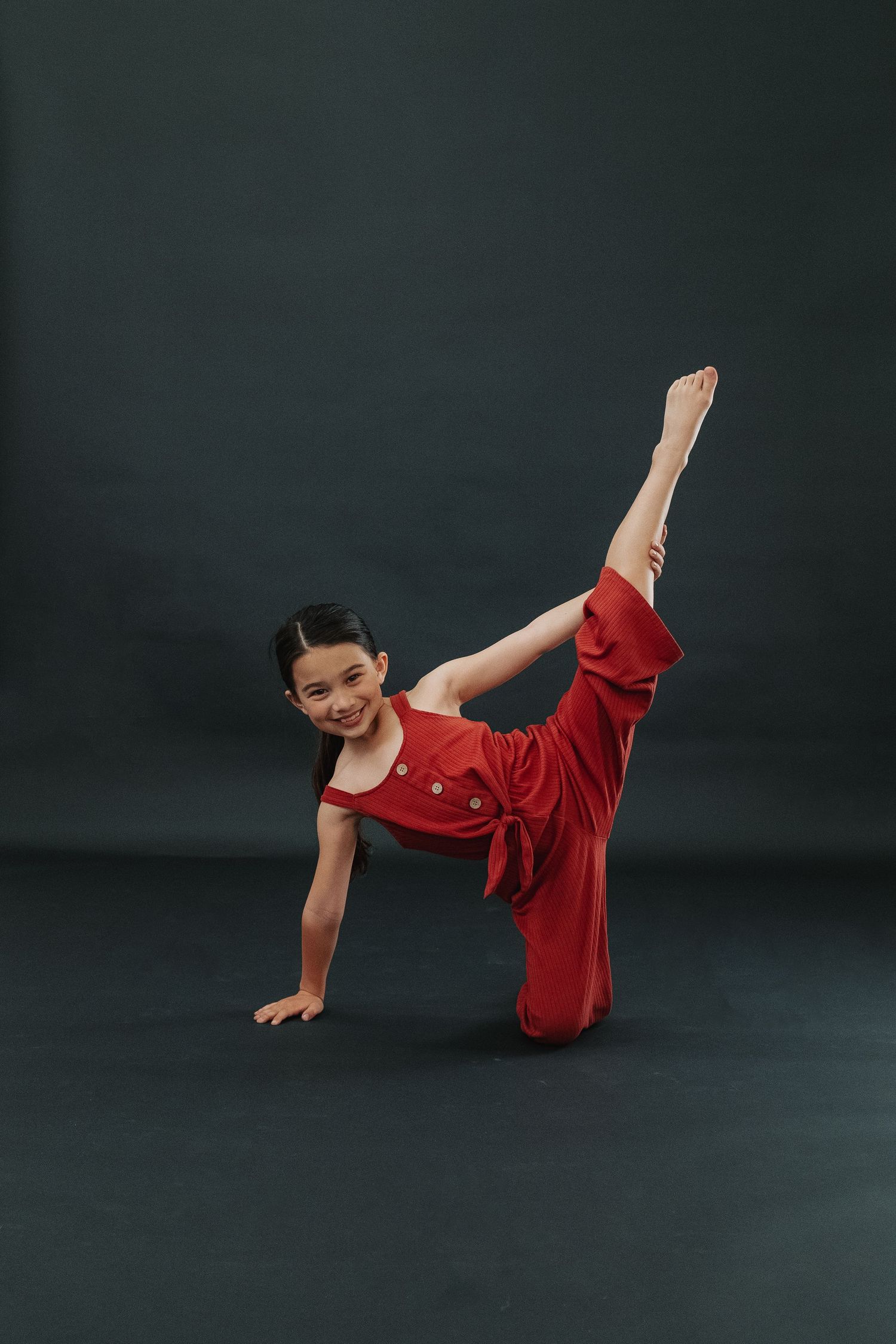 Dancer in red outfit demonstrates high kick pose against dark studio background.