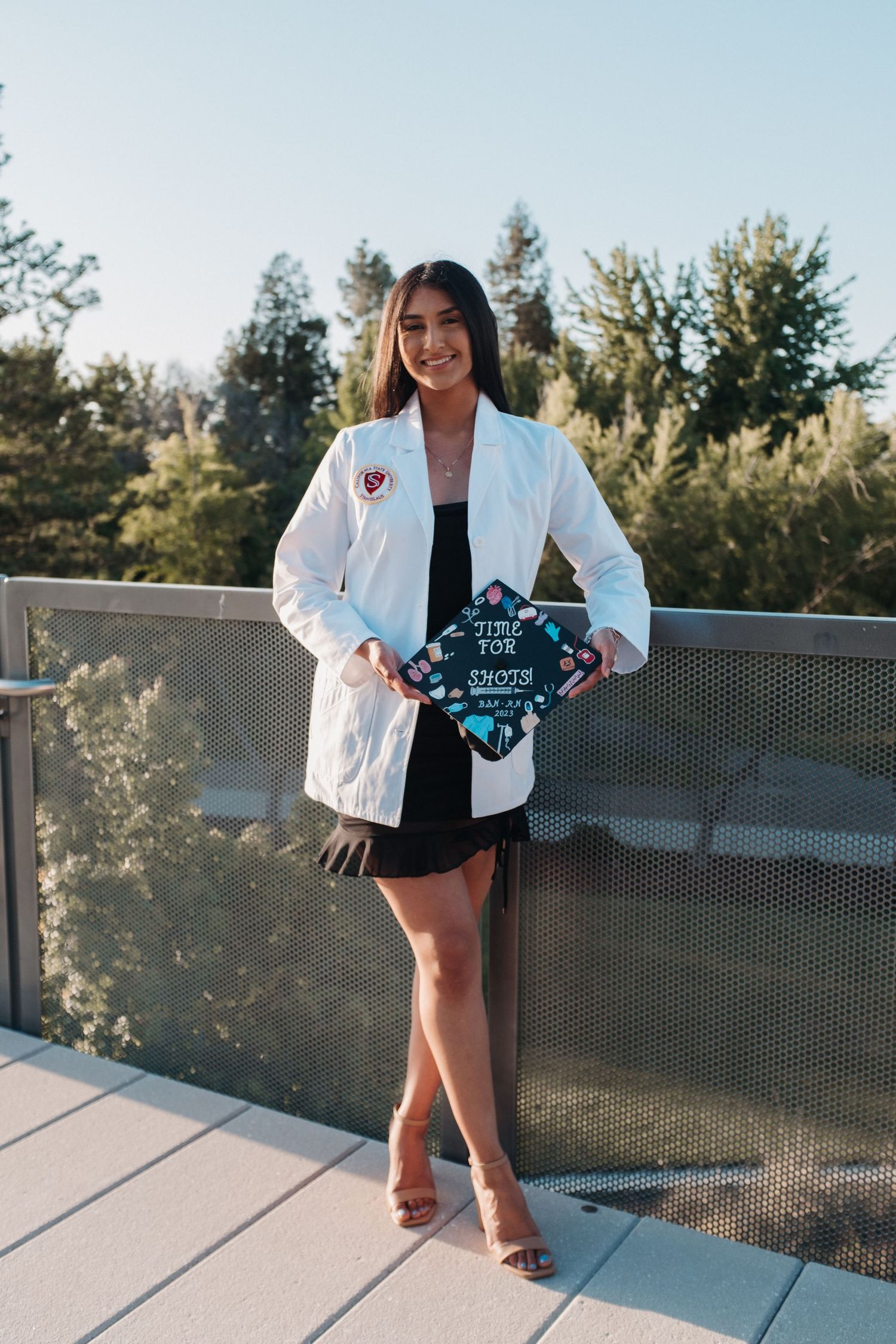 A person in a white lab coat holds a graduation sign while posing on an outdoor balcony.