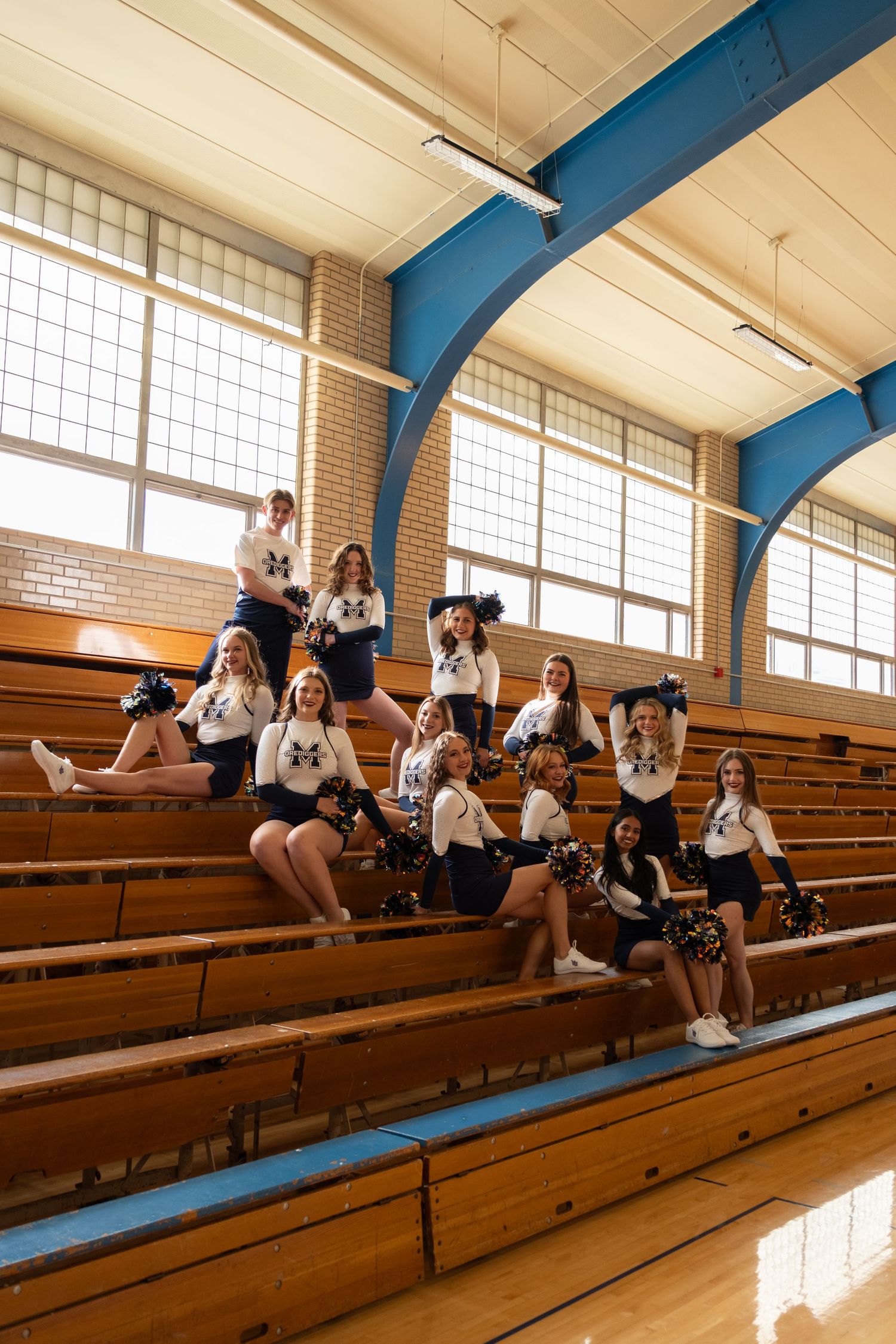 Cheerleading squad posing in school gym bleachers wearing navy and white uniforms with pom-poms.