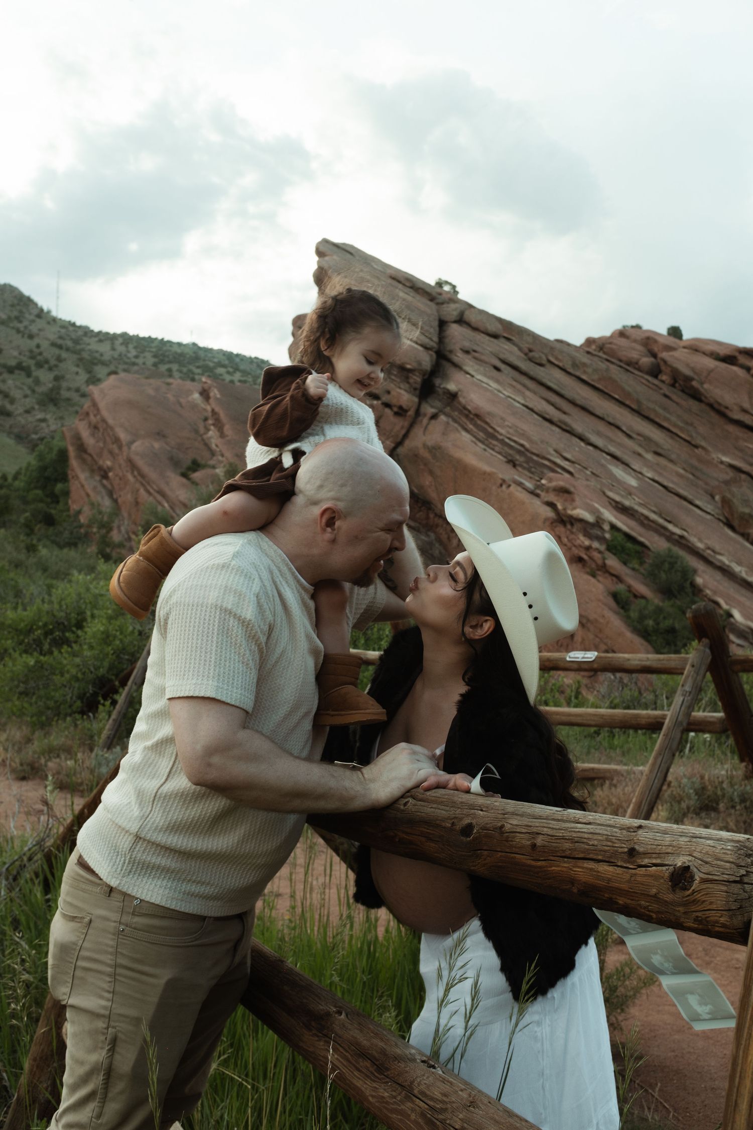Family of three sharing a tender moment at Red Rocks Park, Colorado, with iconic red rock formations in the background.