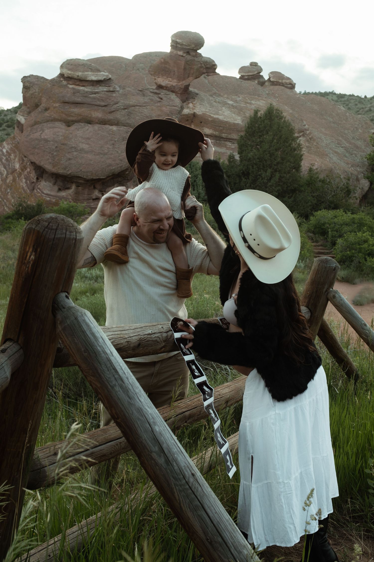 Family with child on shoulders near rustic fence and red rock formations in a scenic outdoor setting.