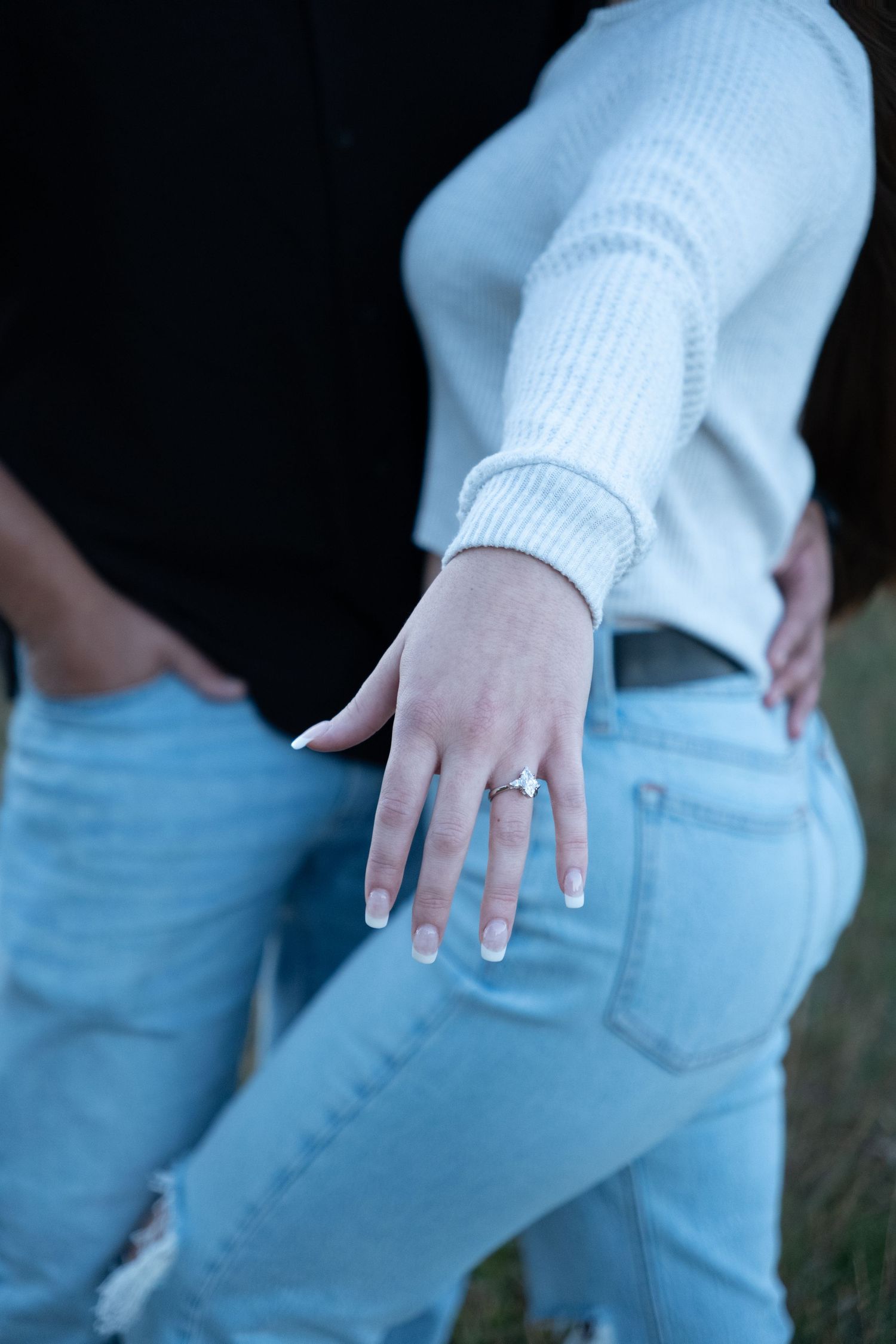 Close-up of a woman's hand displaying an engagement ring while wearing a white sweater and light blue jeans.