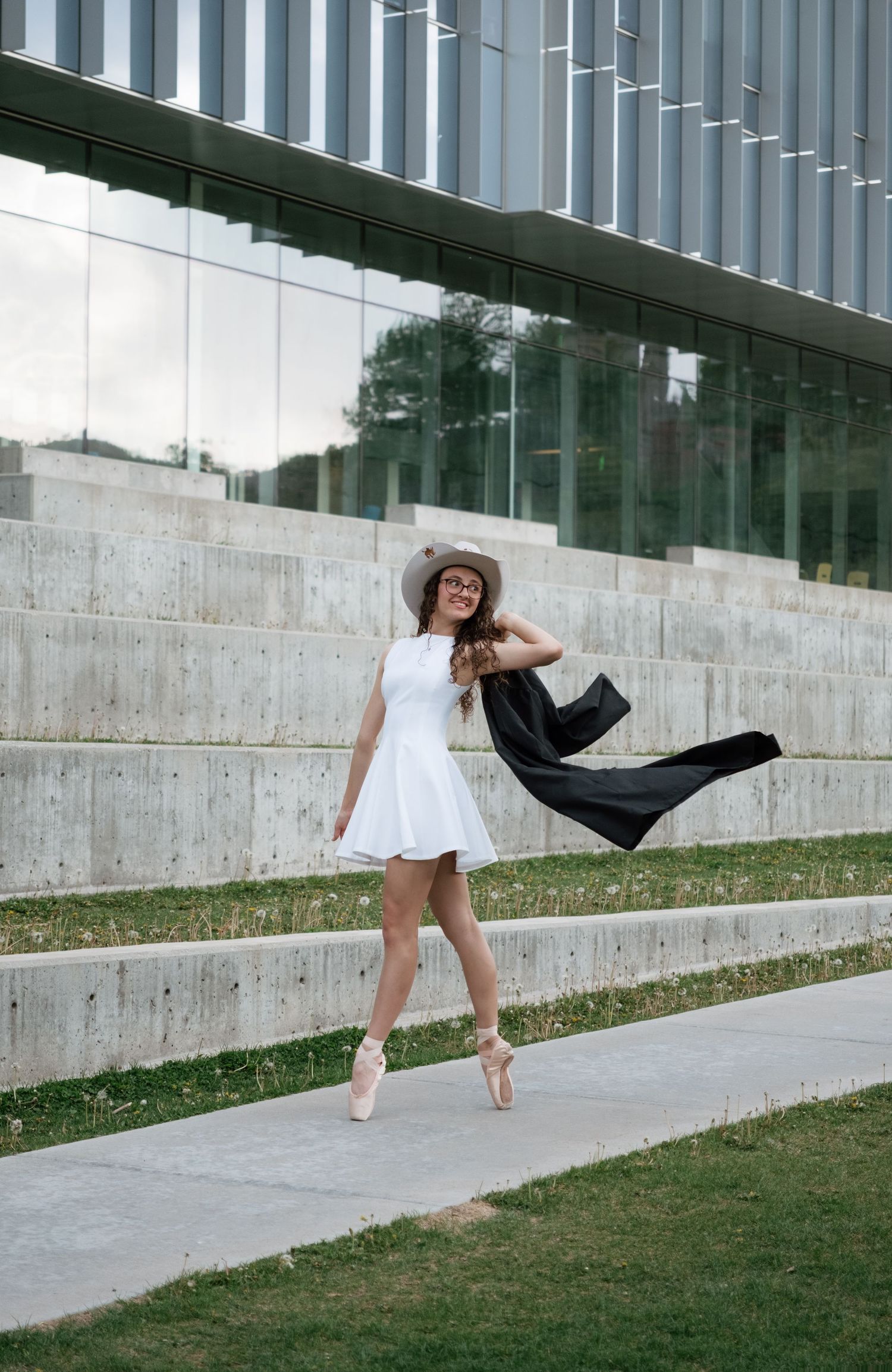 A ballerina in a white dress and hat poses gracefully on pointe shoes outdoors near a modern concrete building.