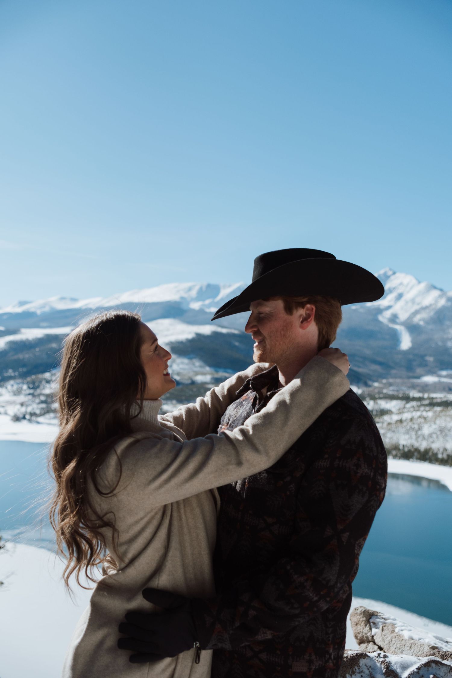 Couple embracing on snowy mountain overlook, man in cowboy hat, winter lake backdrop, blue sky.
