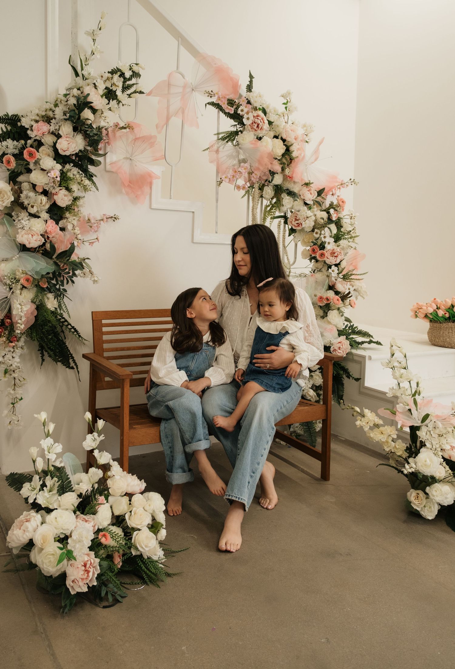 A mother and two young children sit on a wooden bench surrounded by a floral arch of pink and white blooms.