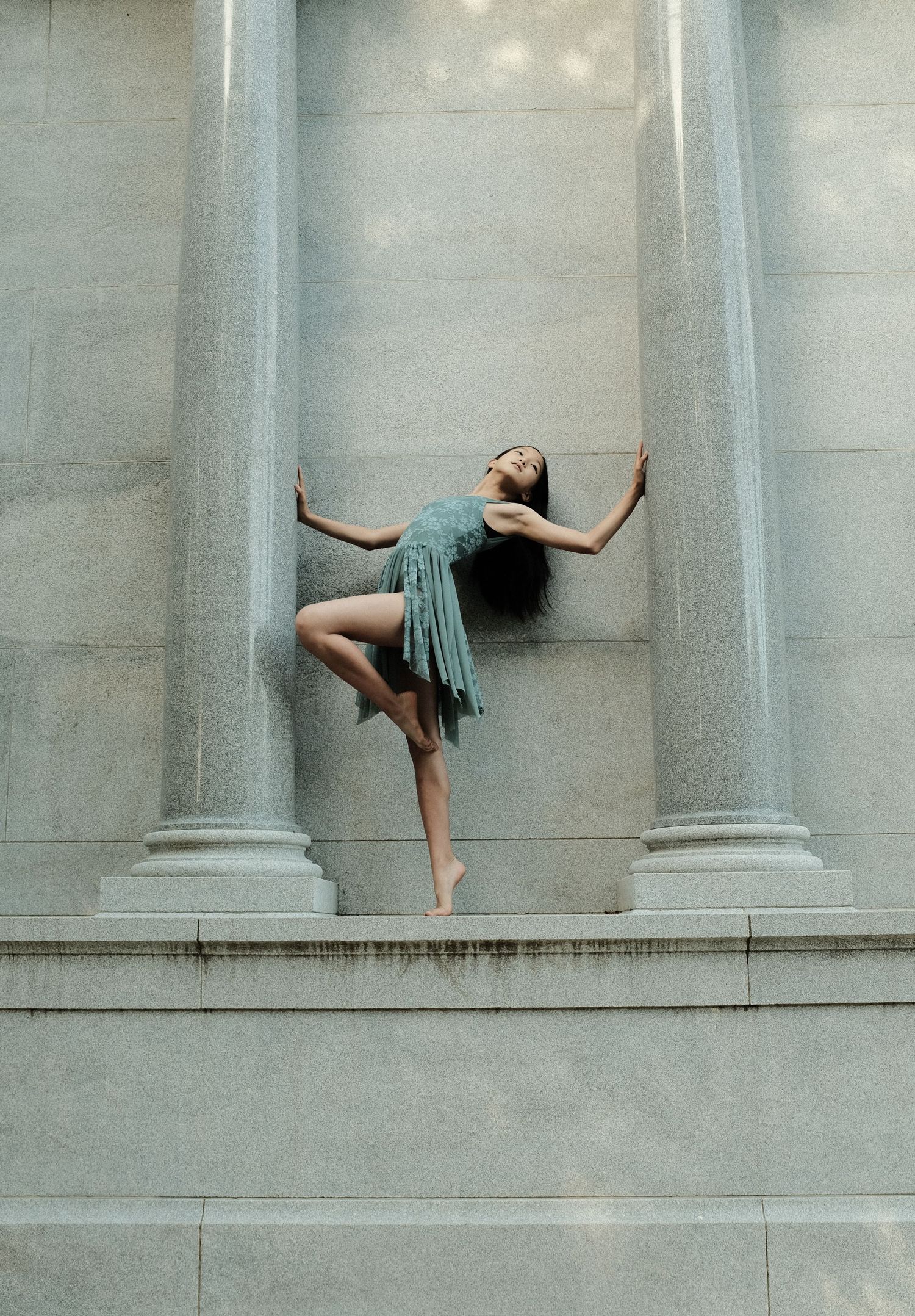 Dancer in teal dress performs artistic pose between classical stone columns.