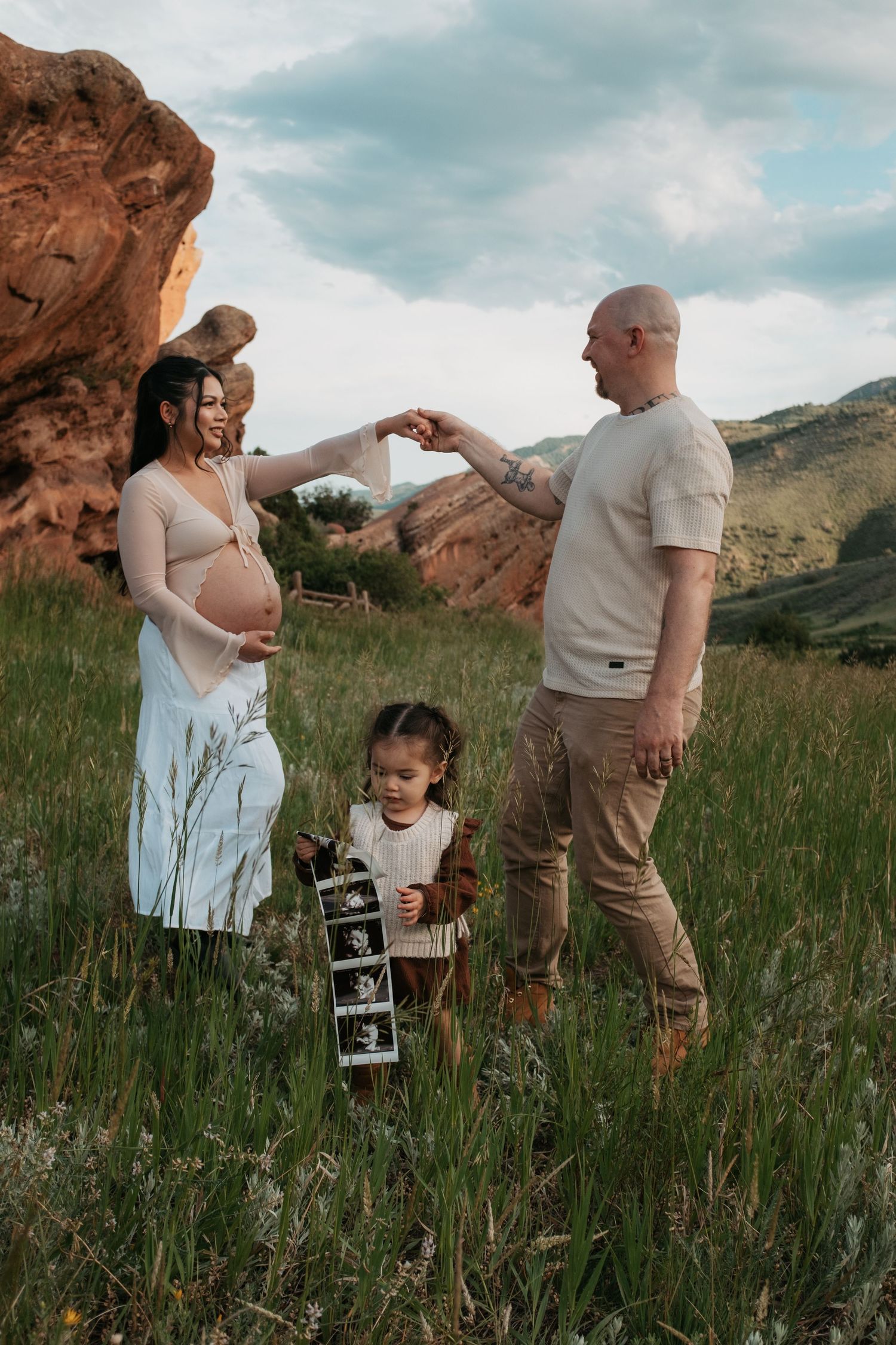 A maternity photo session captures a moment between expectant parents holding ultrasound images outdoors.