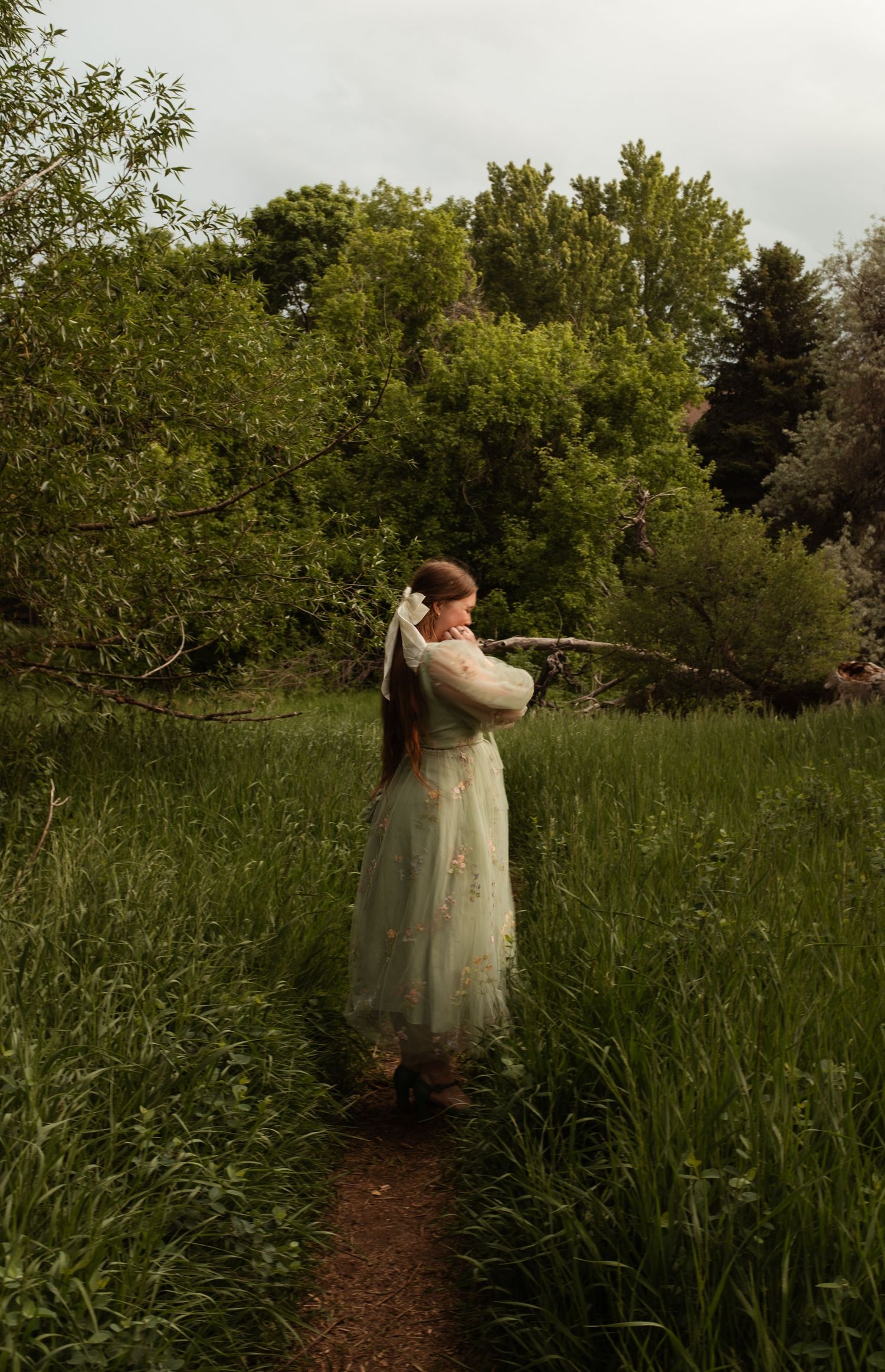 Person in elegant dress poses with extended arms on dirt path through wild meadow.