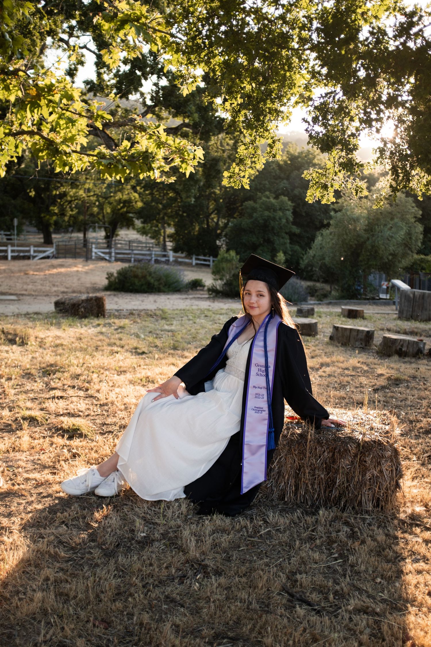 Graduate in academic dress poses on hay bale in sunlit outdoor setting.