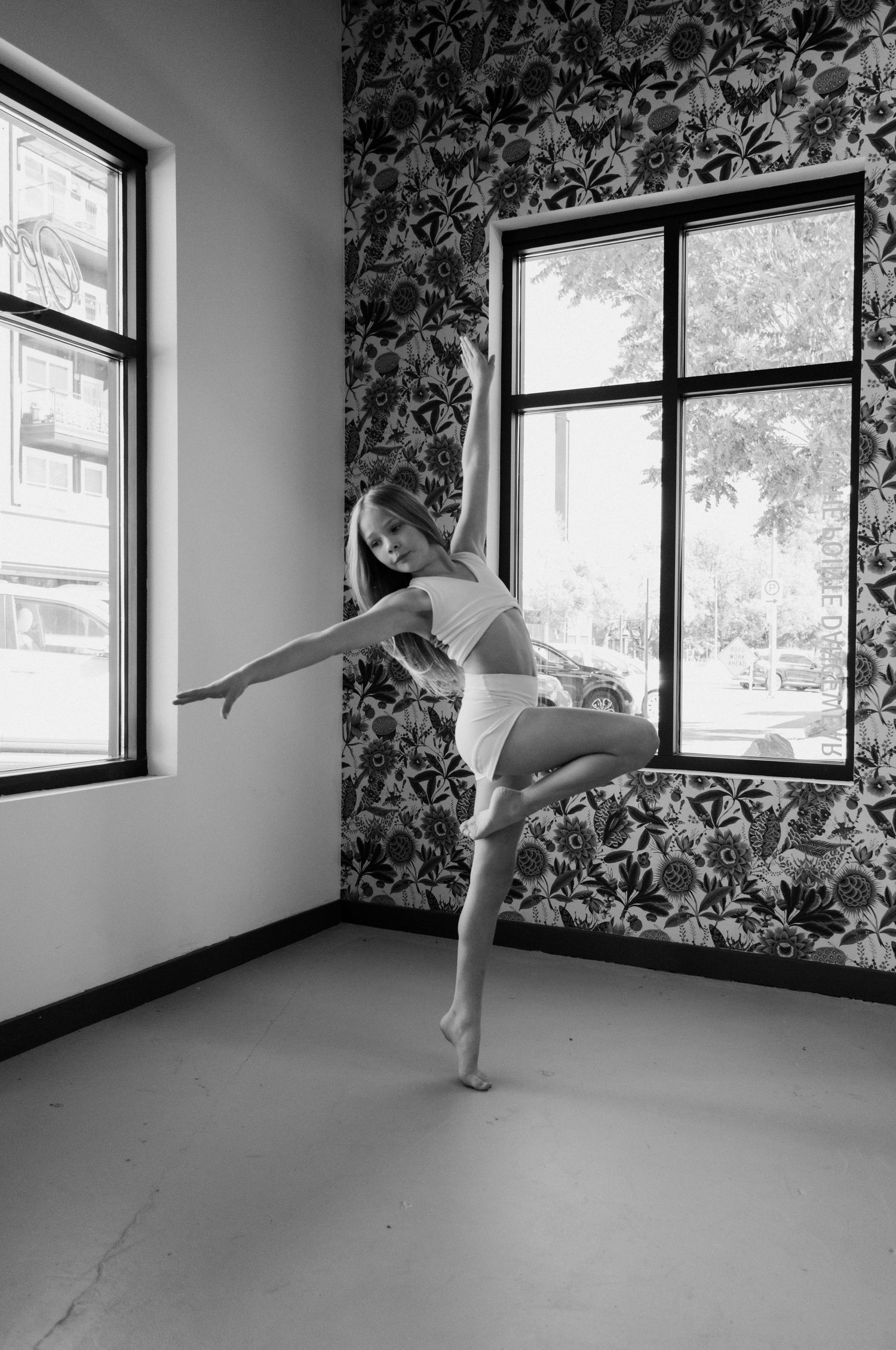 Black and white photo of a young dancer balancing on one foot in an elegant pose inside a floral wallpaper studio.