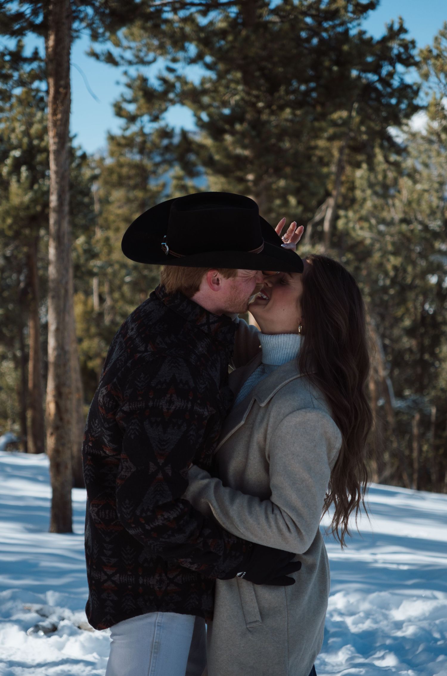 Couple kissing in snowy forest, man wearing black cowboy hat and floral jacket, woman in gray turtleneck sweater.