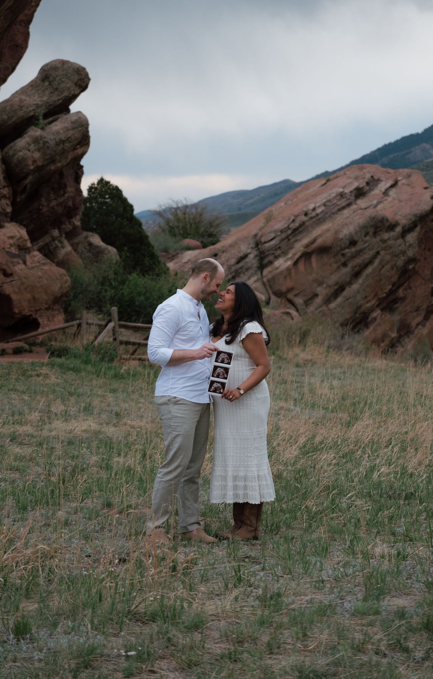 Couple sharing a romantic moment holding ultrasound photos at Red Rocks Park, Colorado, with dramatic red rock formations.