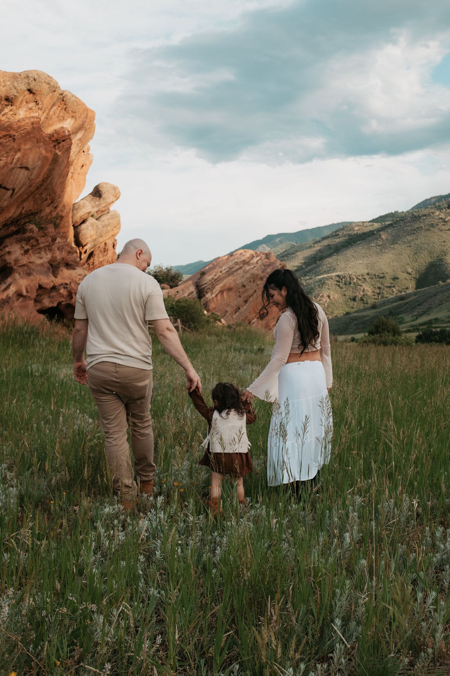 A family of three holds hands walking through a green meadow with red rock formations and mountains in the background.