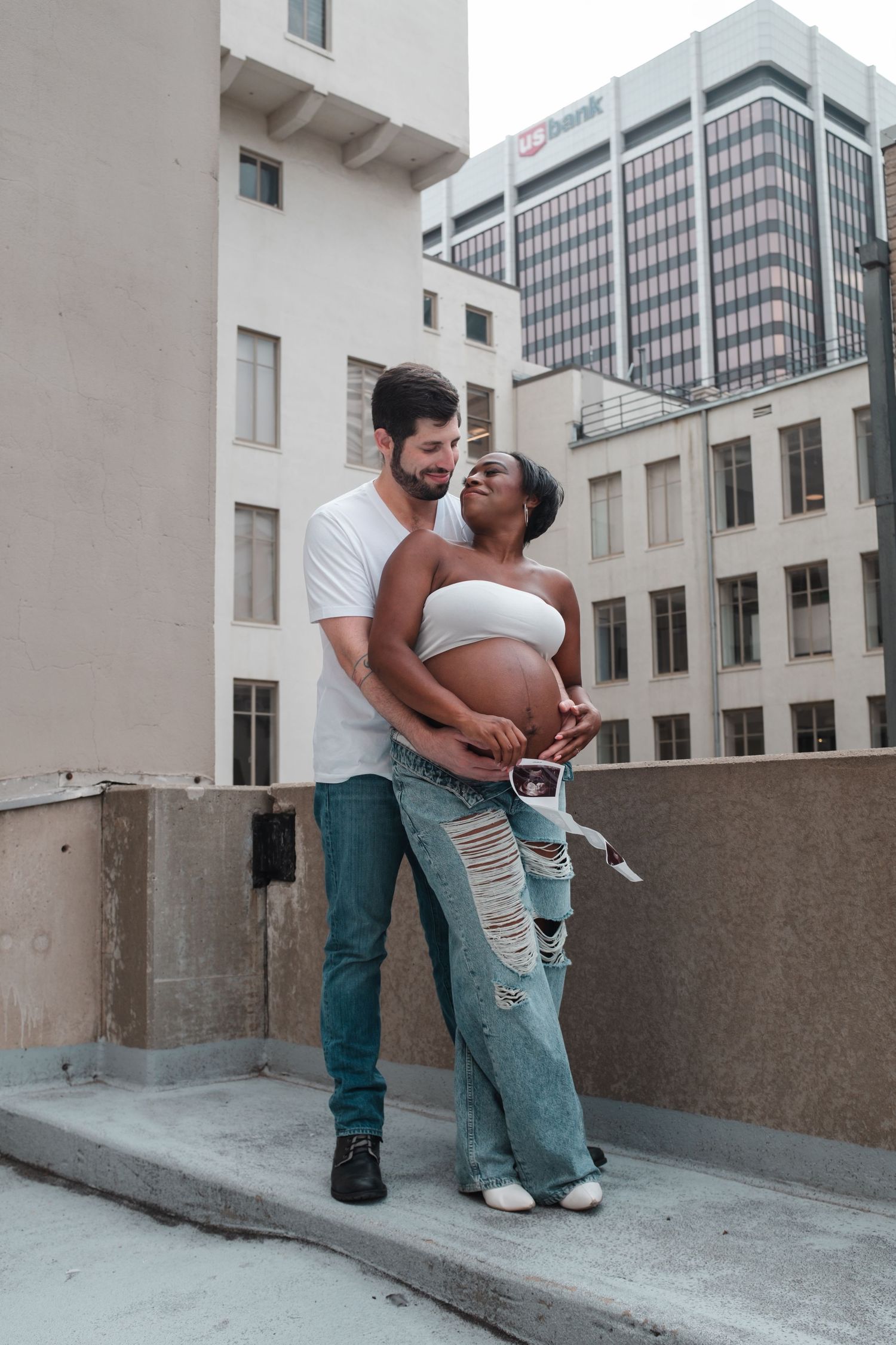 Interracial couple sharing a tender moment on a rooftop, woman visibly pregnant holding ultrasound photos.